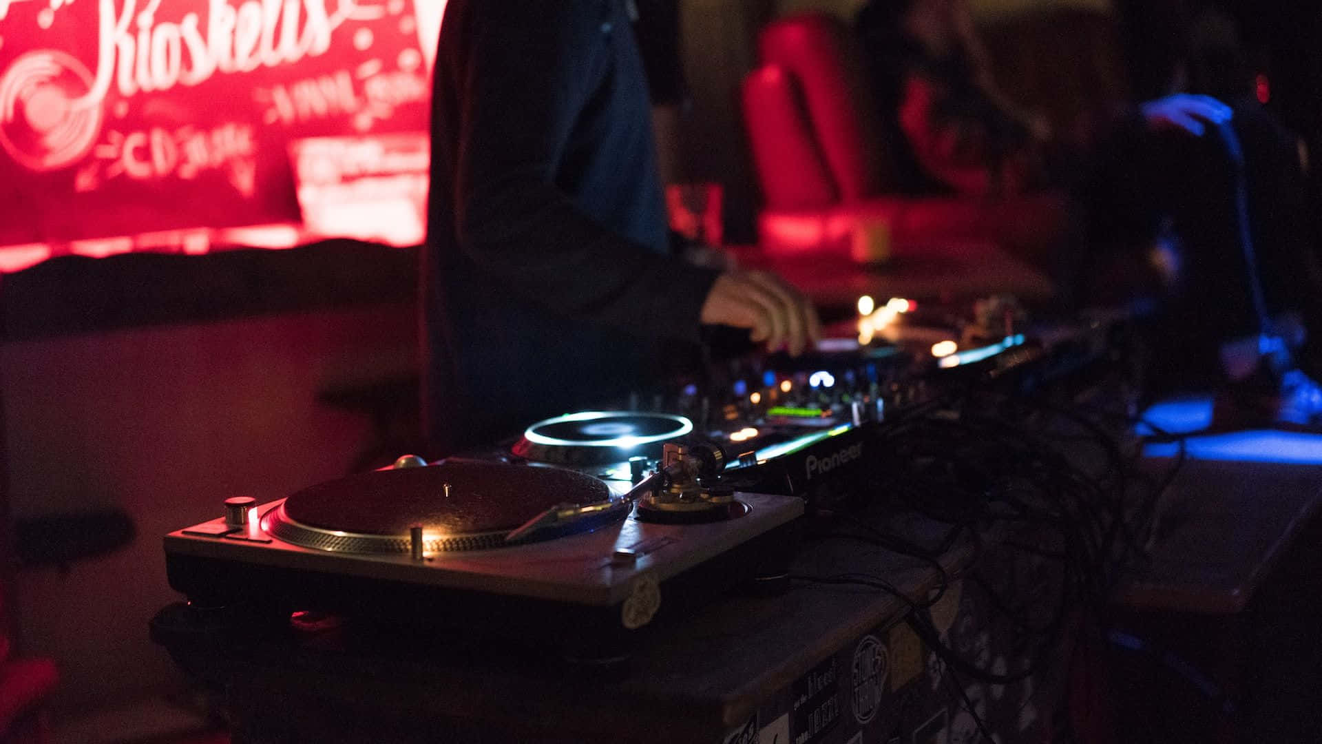 DJ 桌布 壁紙 — DJ 在燈光昏暗的俱樂部中混音，紅色霓虹燈標誌和發光的唱盤投射出色彩繽紛的倒影。