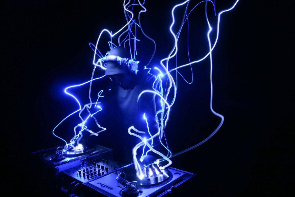 DJ 混音酷炫音樂 壁紙 — 一個剪影中的DJ正在操作唱盤，發光的藍色光軌在黑暗的俱樂部環境中圍繞他們旋轉。