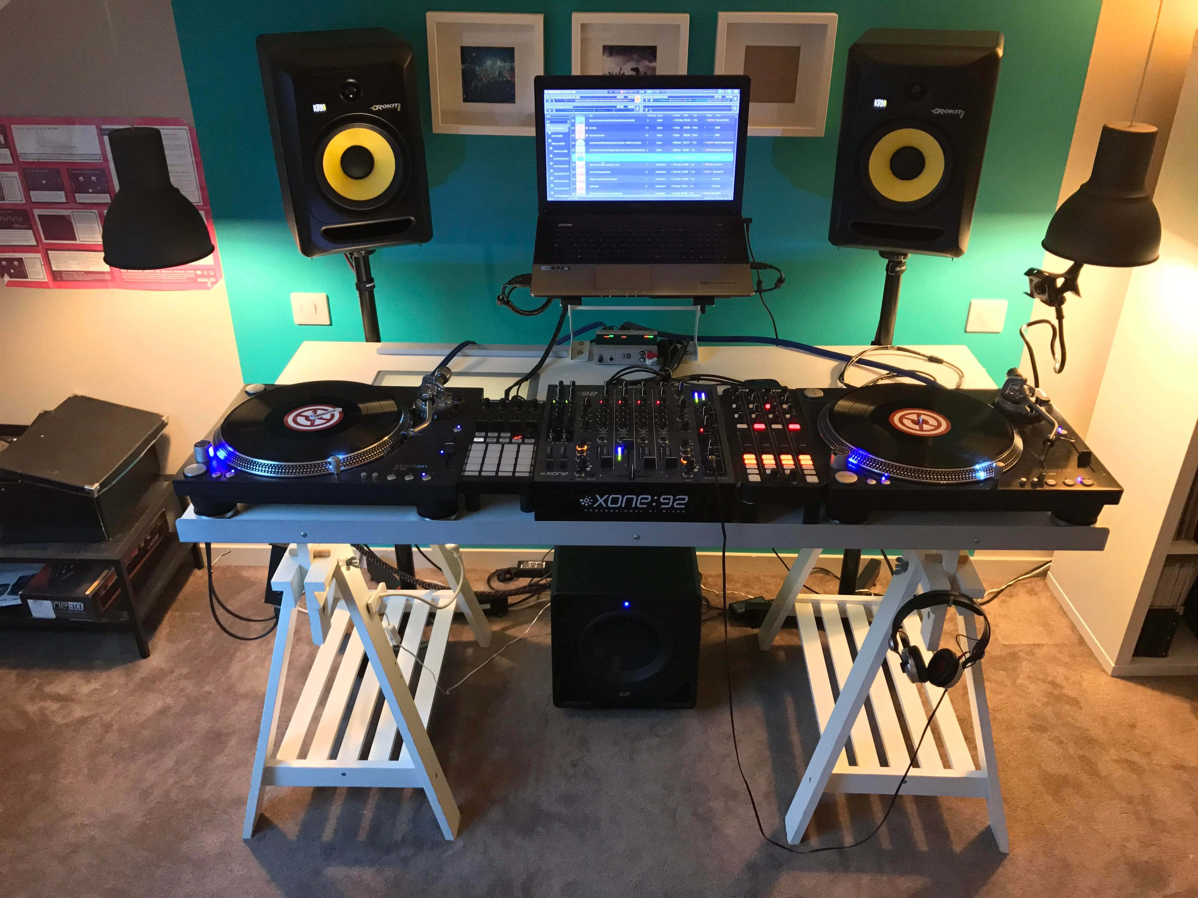 DJ音樂工作室監視器與音箱設置圖 壁紙 — 一個DJ設置，包括兩個唱盤、一個混音器、手提電腦和揚聲器，排列在白色桌子上，背景是青綠色牆壁。