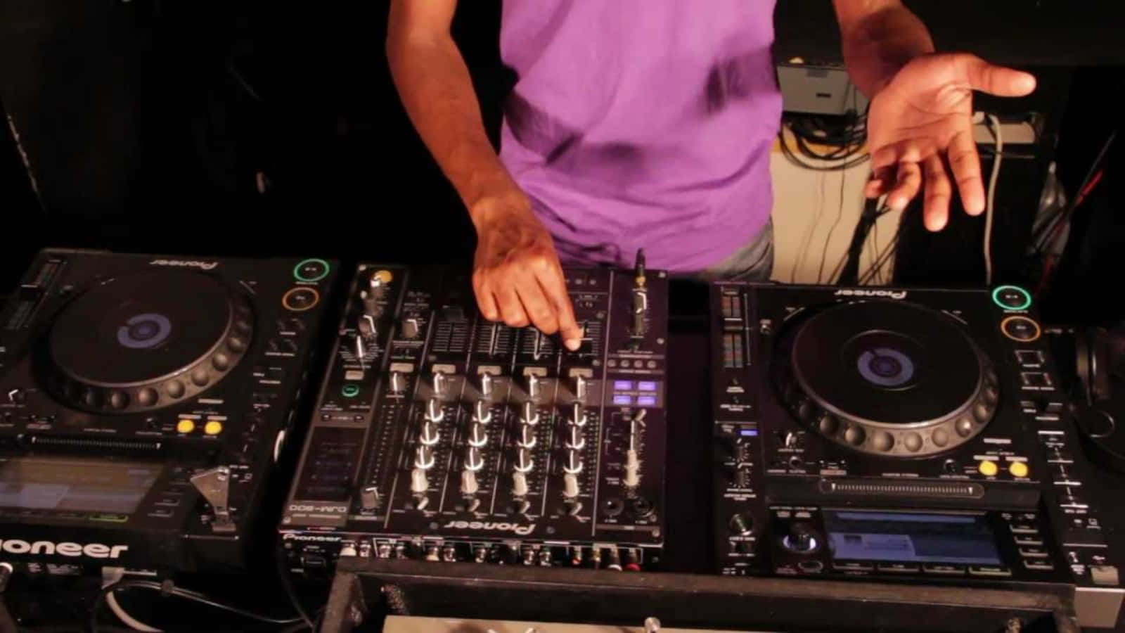 DJ音樂混音器設置圖 壁紙 — 一位身穿紫色襯衫的DJ在昏暗的舞台燈光下，於黑色Pioneer唱盤上混音曲目。