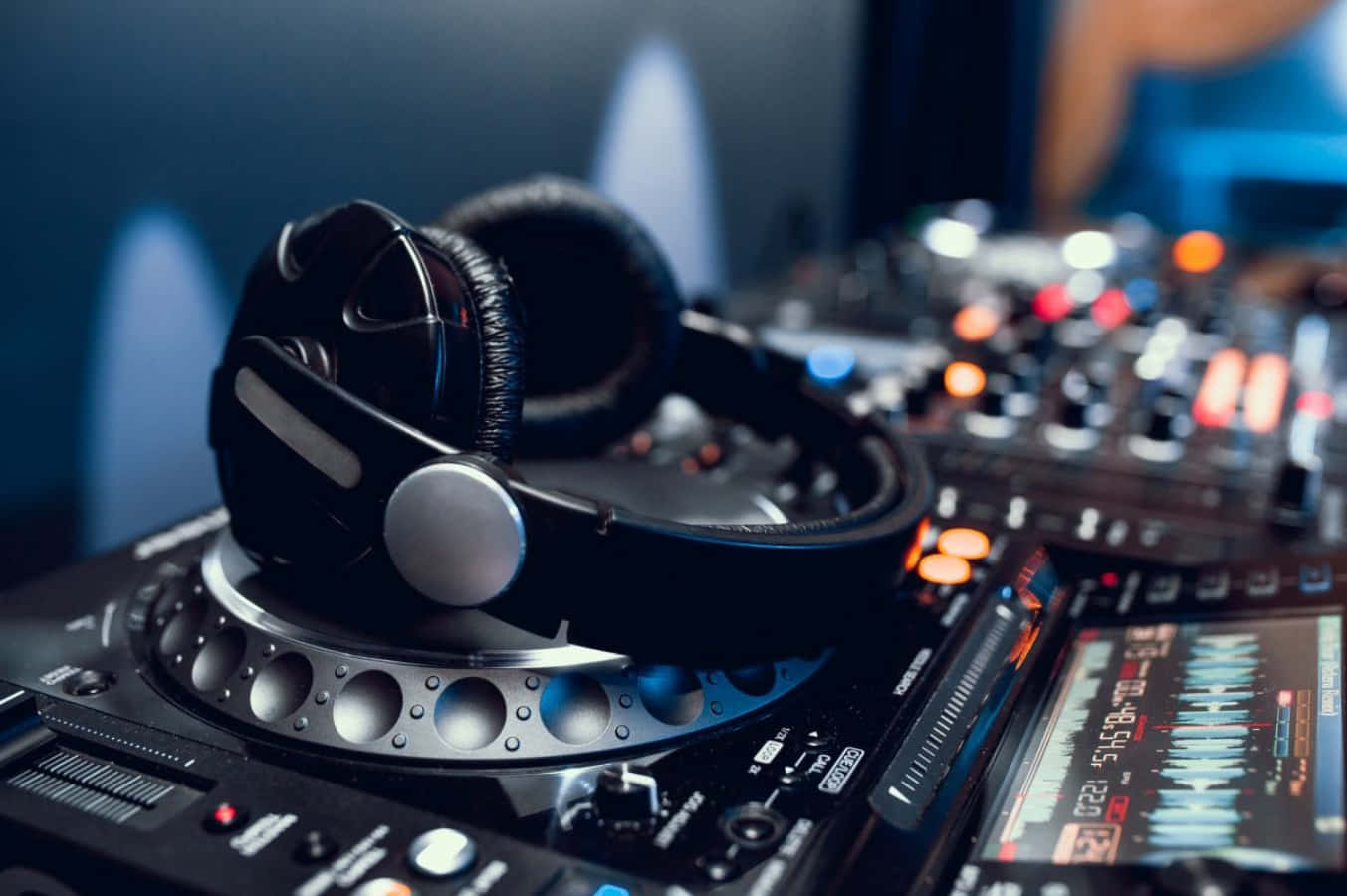 DJ音樂設置搭配耳機圖片 壁紙 — 專業DJ耳機放在帶有發光控制裝置和波形顯示器的唱盤上，身處燈光昏暗的俱樂部。