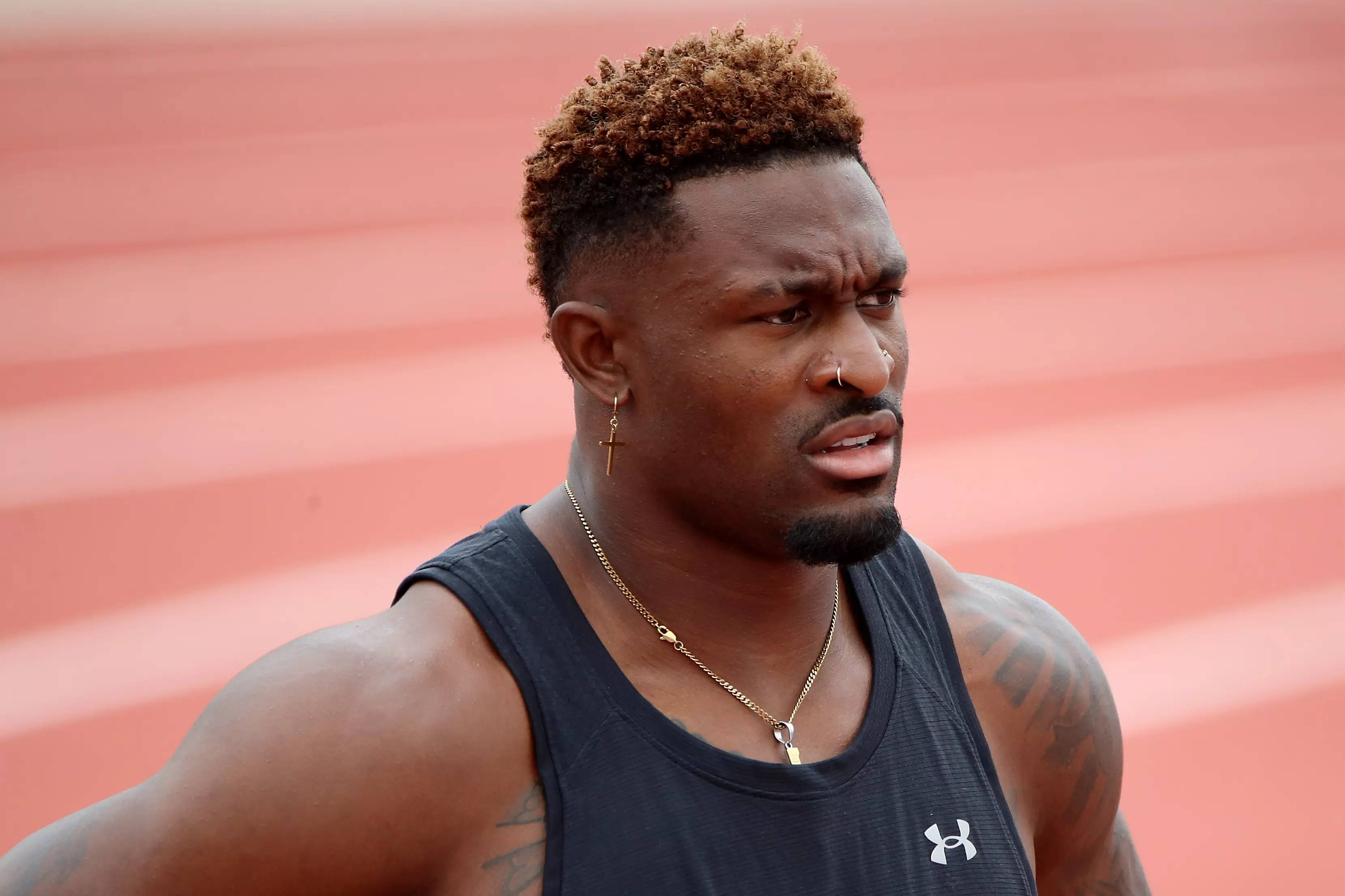 DK Metcalf 黑色背心造型 壁紙 — 一位專注的男性運動員，捲髮、有紋身，身穿黑色背心站在紅色跑道上。