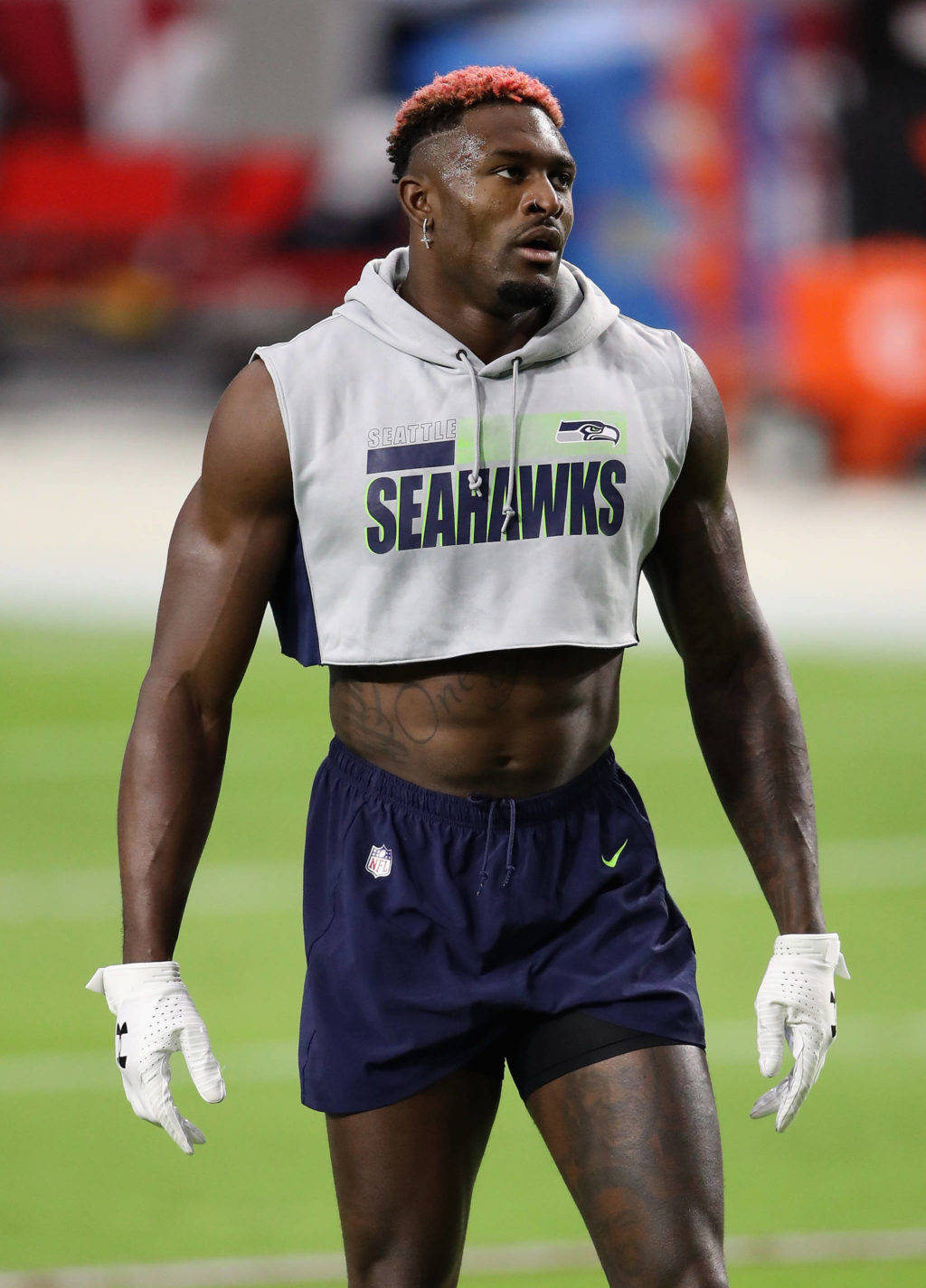 DK Metcalf 雄壯體格 壁紙 — 一位西雅圖海鷹隊美式足球員，頭髮尖端染紅，身穿短版連帽衫和海軍藍短褲，站在綠色球場上。