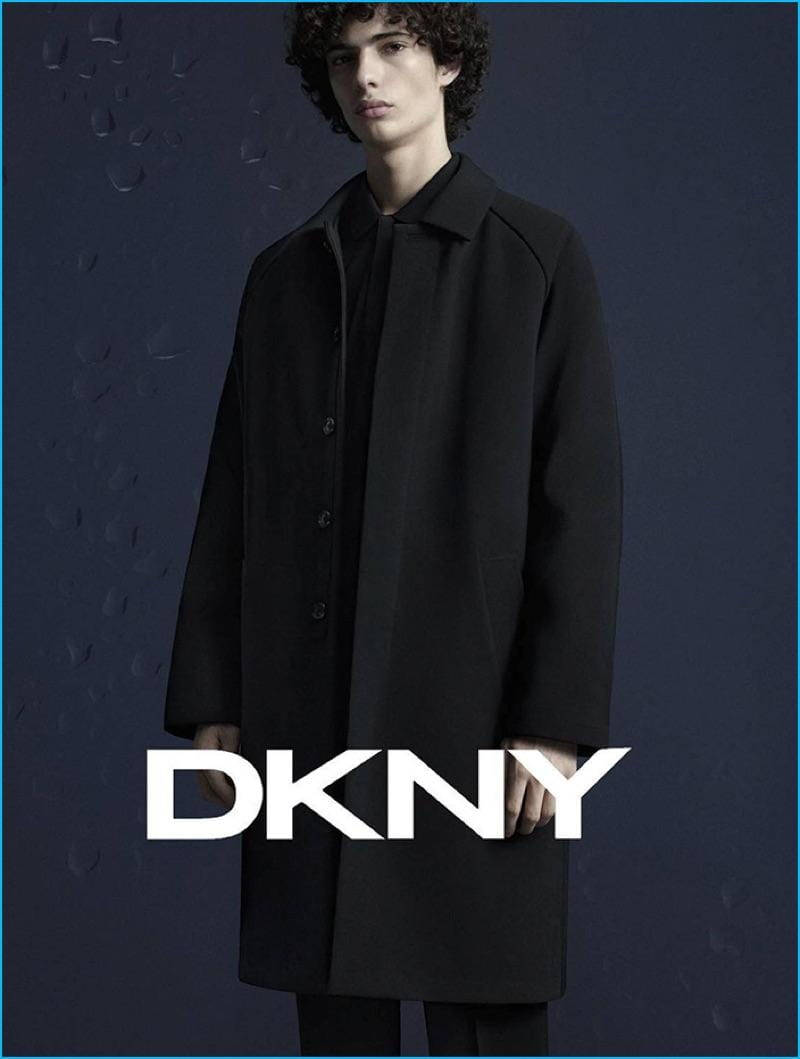DKNY促銷海報 壁紙 — 一位捲髮年輕男子穿著時尚黑色外套，背景是帶有水珠和醒目DKNY標誌的深藍色紋理牆壁。