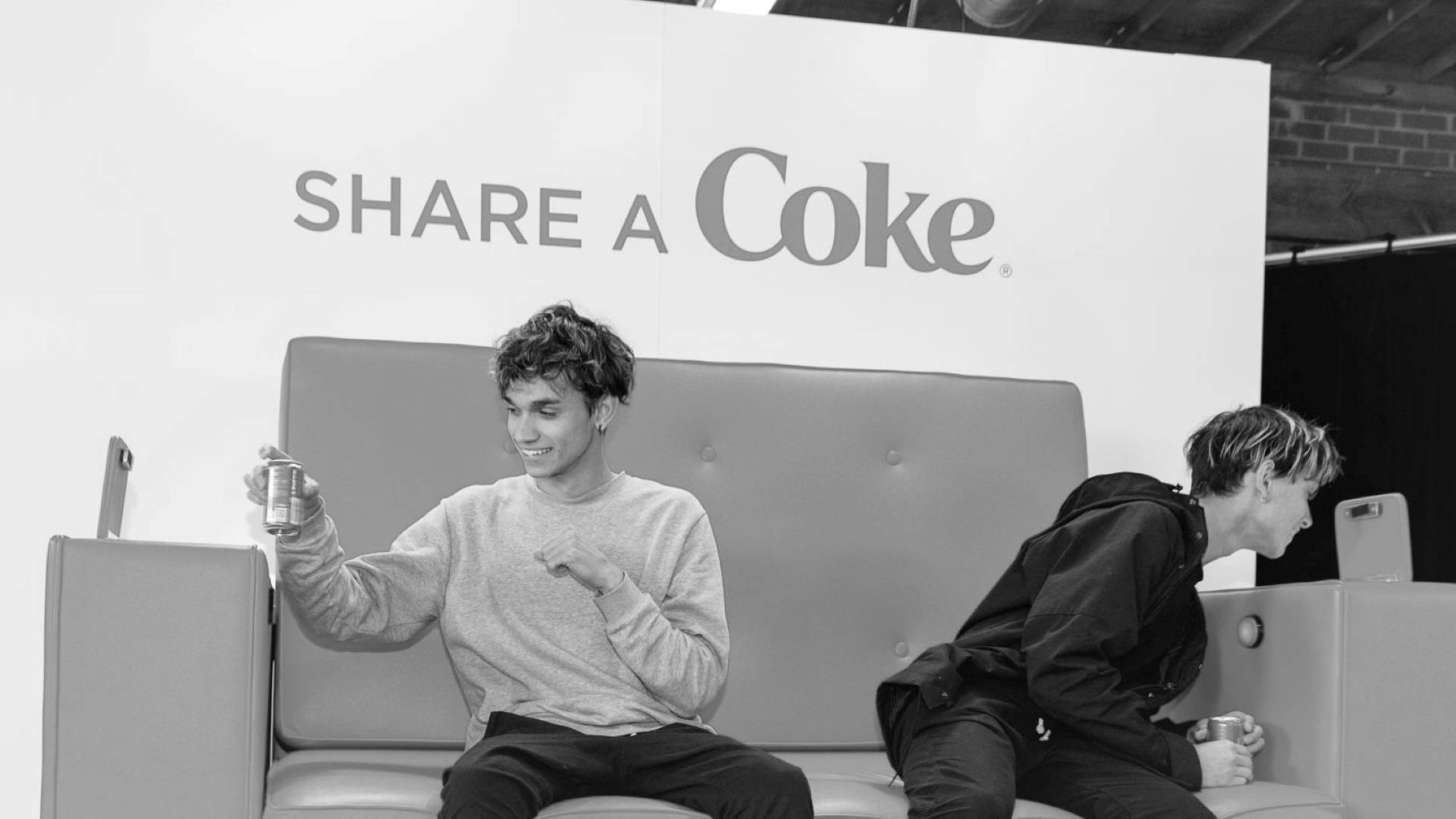 兩名年輕男子坐在沙發上微笑，手持可口可樂罐，身後有Share a Coke品牌標誌。