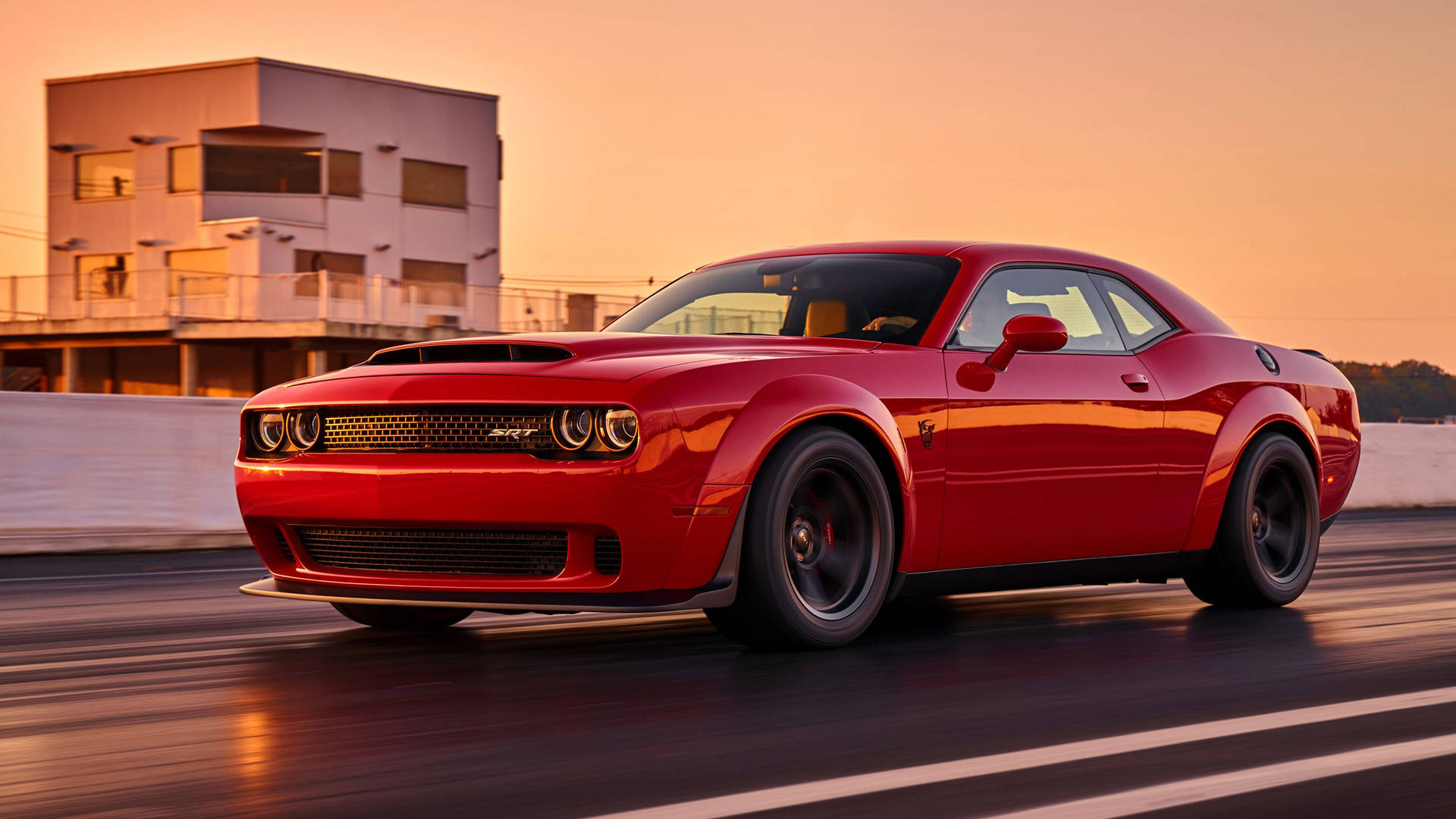 紅色 Dodge Challenger 在日落時分的賽道上飛馳，後面有一座現代建築。