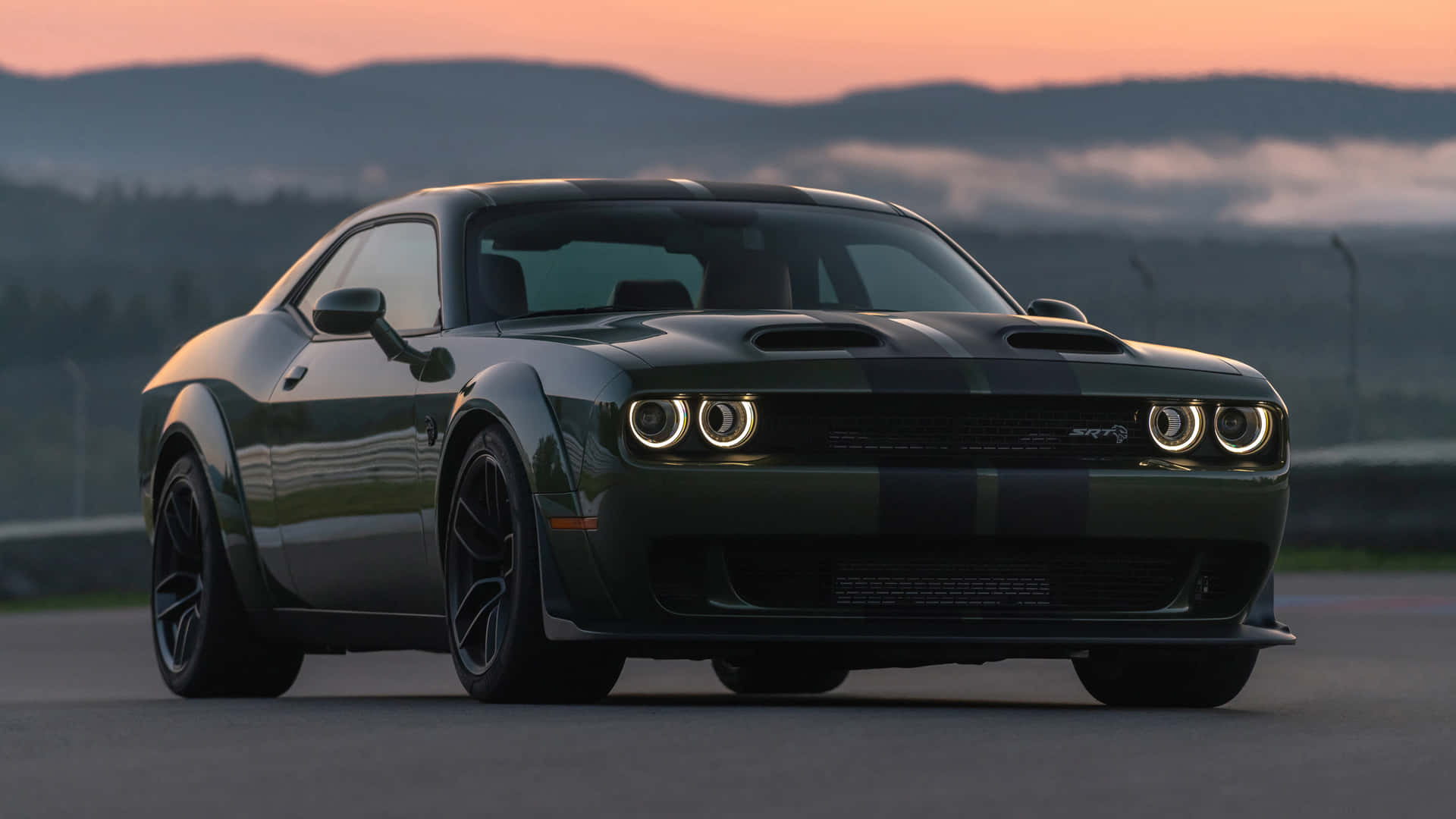 日落時分，深綠色 Dodge Challenger SRT Hellcat 在賽道上，背後有山脈和霧氣。