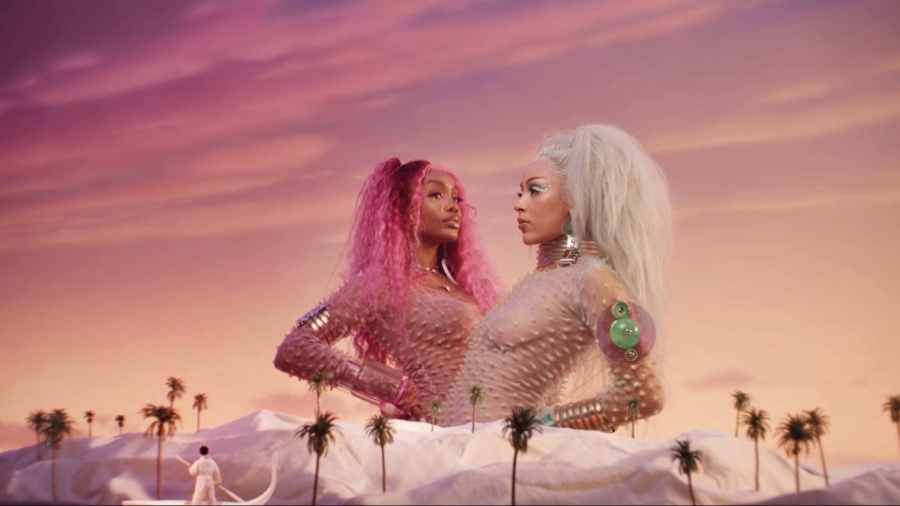 Doja Cat《Kiss Me More》夢幻科幻場景 壁紙 — 兩位粉紅和白色頭髮的女性站在超現實的沙漠景觀中，粉紅色的天空下有棕櫚樹。