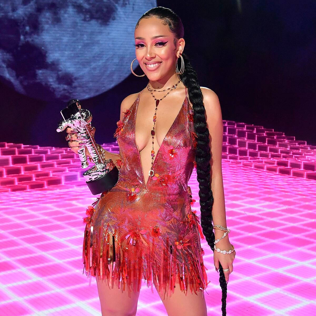 Doja Cat 太空人 壁紙 — 一位微笑的女子身穿閃亮粉紅色流蘇裙，在充滿活力的粉紅色網格舞台上，以月亮為背景，手持獎盃。