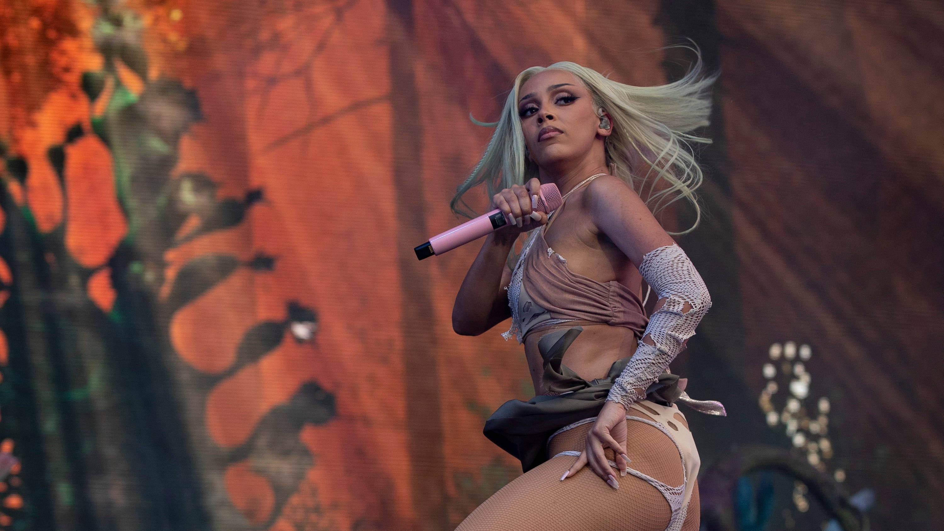 Doja Cat 挥髮造型 壁紙 — 一位金髮歌手在舞台上表演，手持粉紅色麥克風，背景是火熱的橙色。