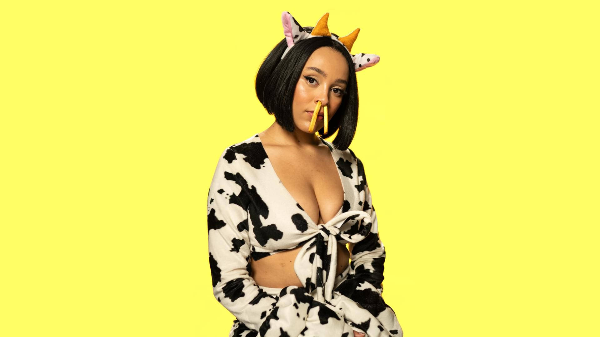 Doja Cat 牛仔服裝造型 壁紙 — 一位身穿乳牛印花服裝和角狀頭帶的女性在亮黃色背景前擺姿勢。