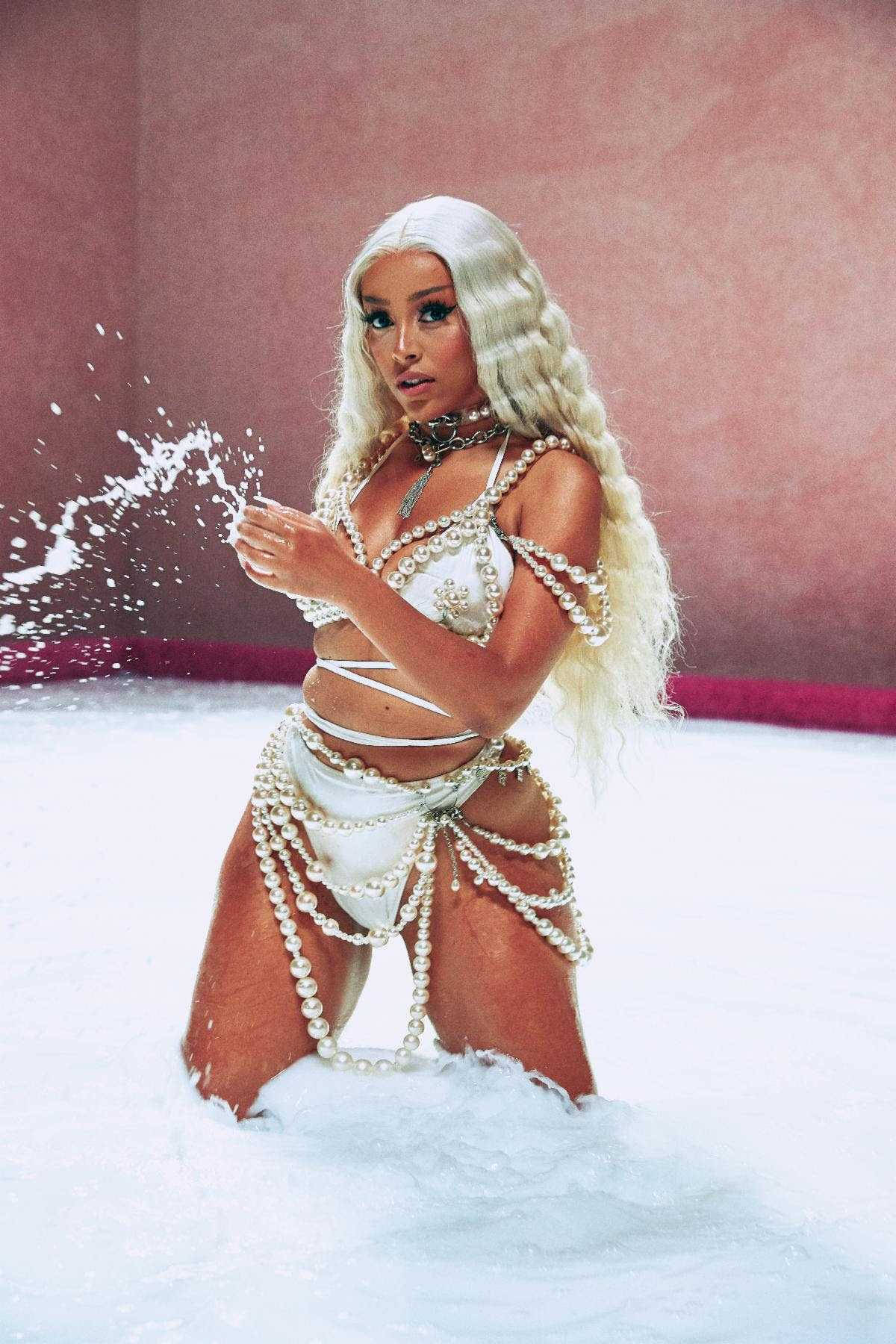 Doja Cat 牛奶浴 壁紙 — 一位留著金色長髮、身穿珍珠裝飾白色服裝的女性，在粉紅色牆壁前的泡沫池中跪著潑水。