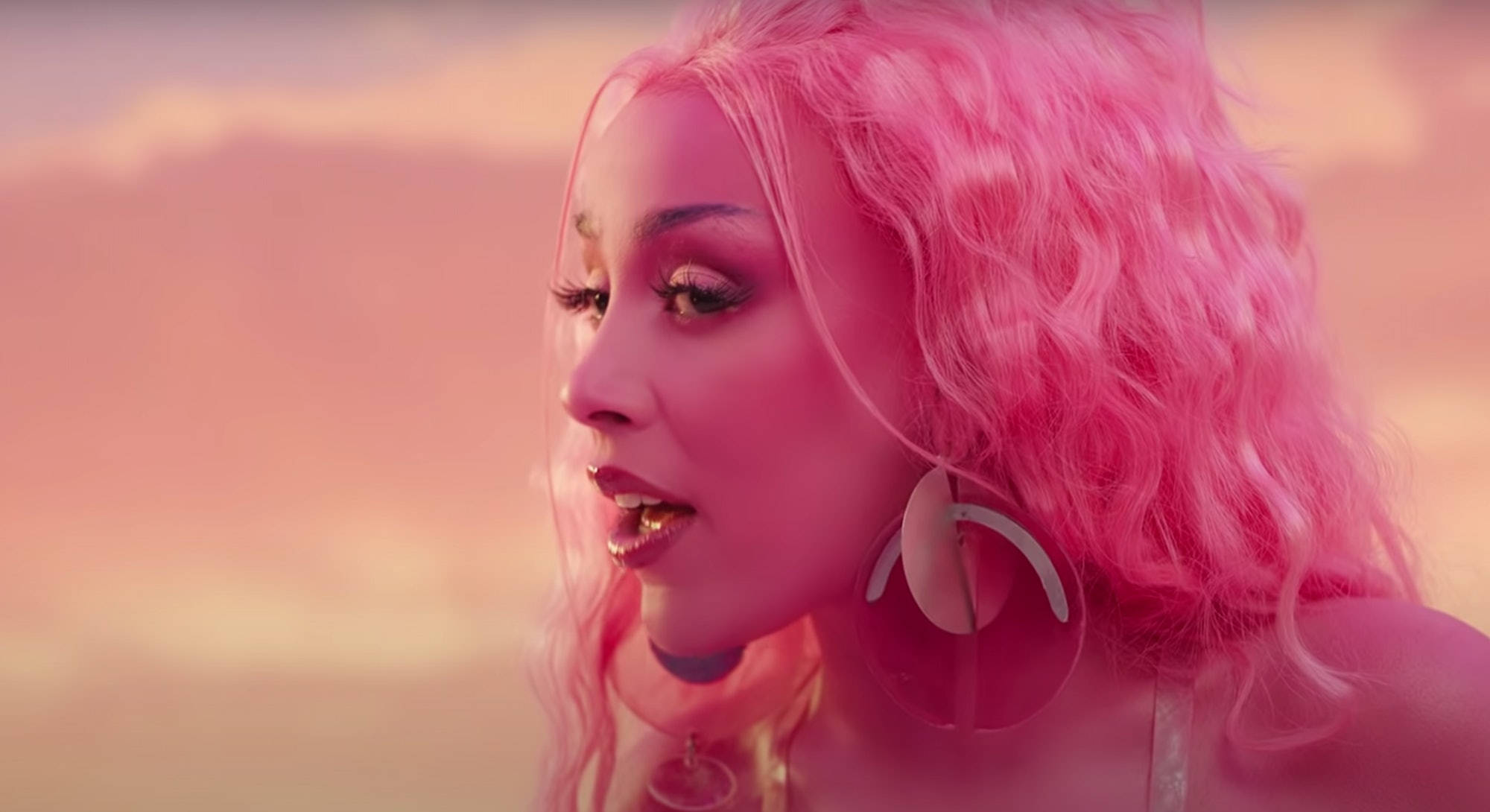 Doja Cat 粉色夢幻風 壁紙 — 一位擁有充滿活力的粉紅色捲髮和大膽妝容的女性，在柔和的粉紅色日落天空下若有所思地凝視。