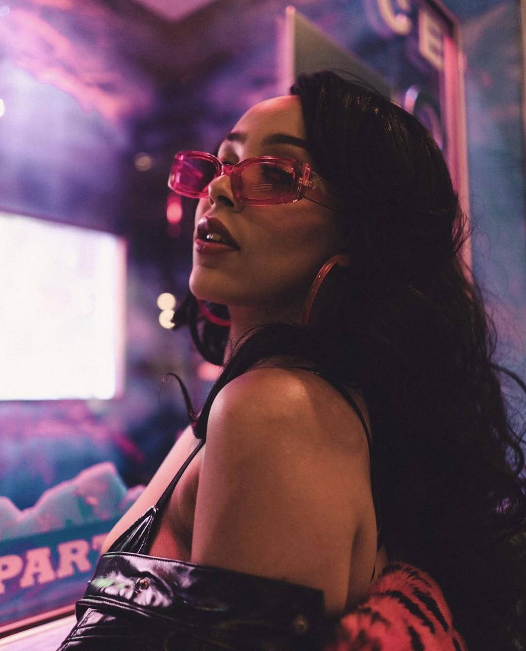 Doja Cat 粉色太陽眼鏡 壁紙 — 一位長黑髮的女性在霓虹燈俱樂部中戴著大膽的粉紅色太陽眼鏡和黑色上衣，周圍有發光的標誌和煙霧。
