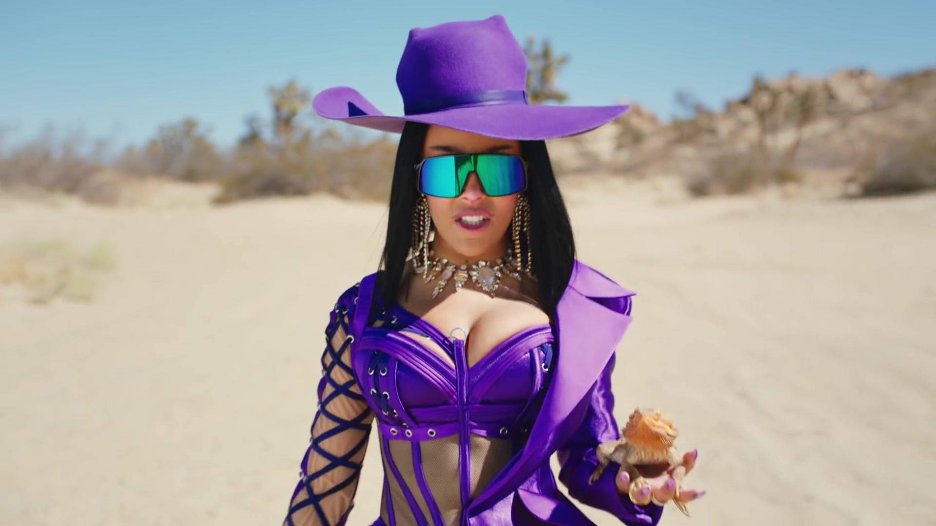 Doja Cat 紫色造型 壁紙 — 一位身穿大膽紫色牛仔裝束和太陽眼鏡的女性，自信地站在陽光普照的沙漠景觀中。