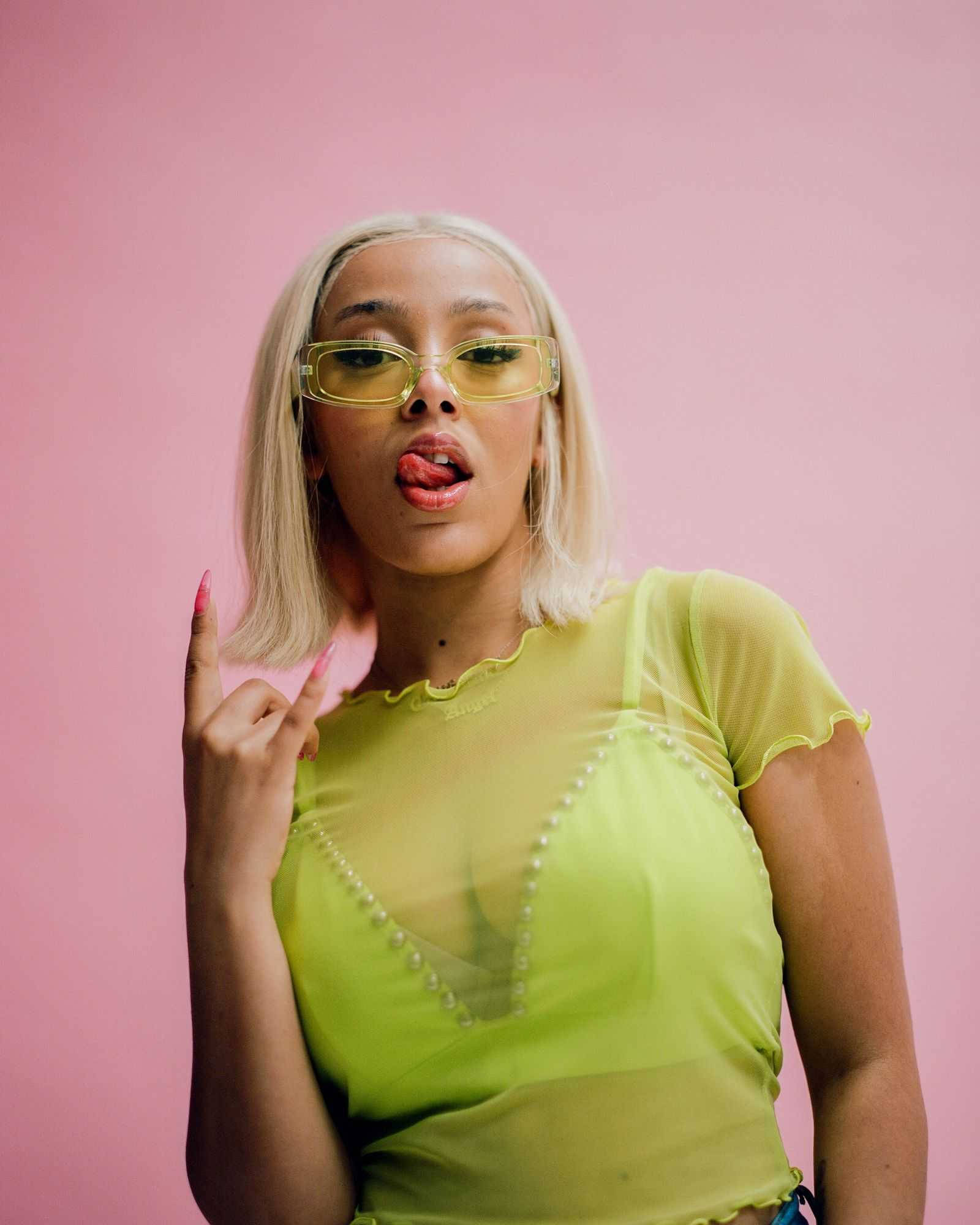 Doja Cat 青檸上衣 壁紙 — 一位金髮、戴黃色眼鏡的女性，在粉紅色背景前伸出舌頭並做出手勢。