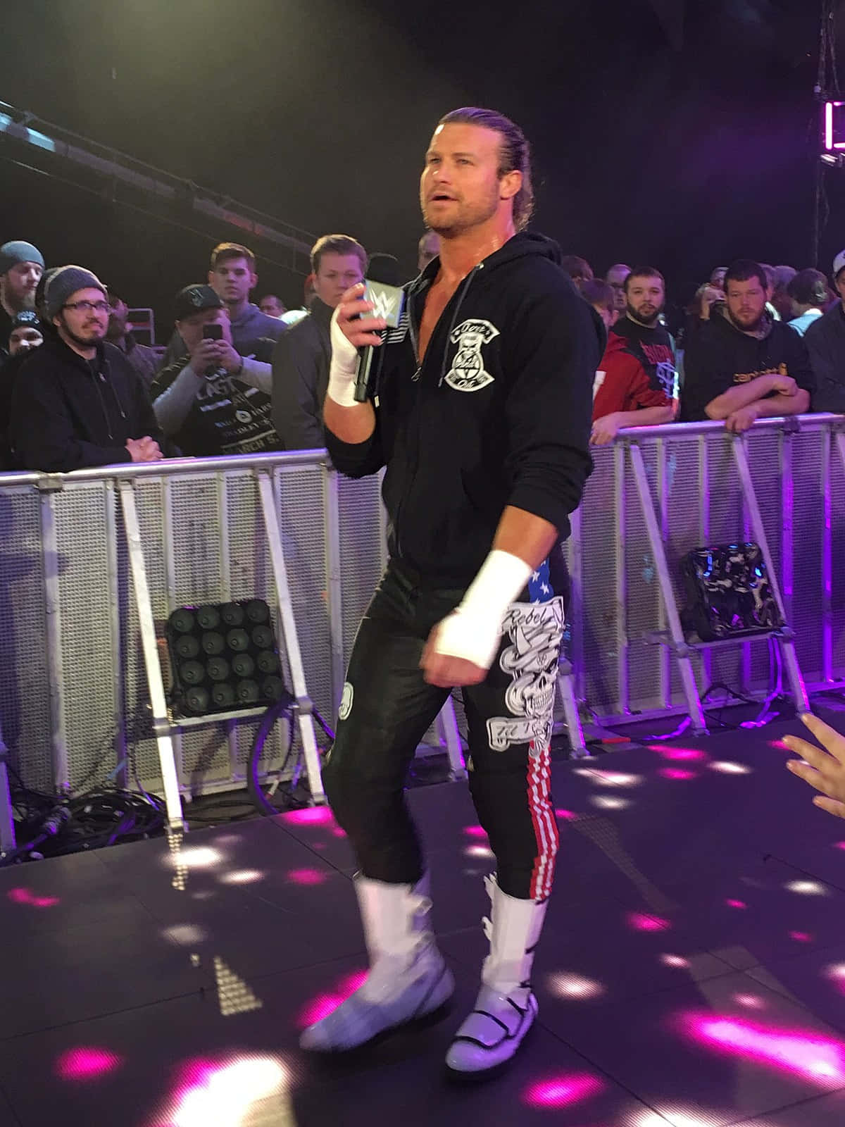 Dolph Ziggler脫口秀喜劇演員 壁紙 — 一名穿著黑色連帽衫和愛國褲子的摔角手站在燈光璀璨的舞台上，手持麥克風，粉絲們在護欄後觀看。