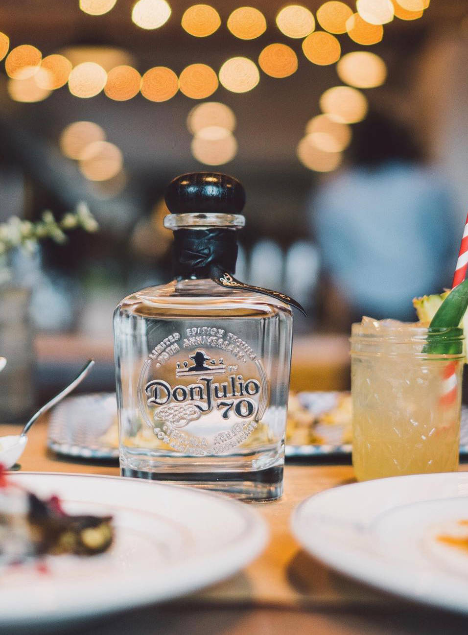 Don Julio龍舌蘭酒空瓶 壁紙 — 一瓶透明的Don Julio 70龍舌蘭酒放在木桌上，旁邊有熱帶雞尾酒和甜點，在溫暖的散景燈光下。