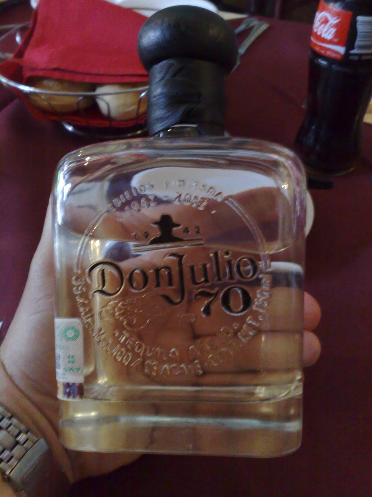 手持Don Julio龍舌蘭酒70度的人 壁紙 — 一隻手拿著透明嘅Don Julio 70龍舌蘭酒樽，黑色樽蓋，紅色枱布上面，背景有可口可樂樽。