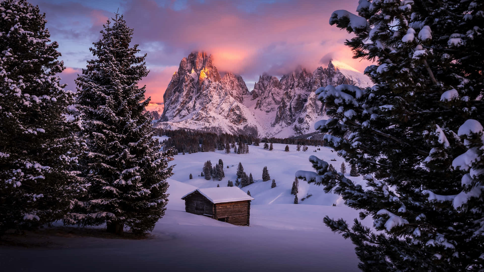  壁紙 — 一座孤獨的雪覆蓋小屋坐落在冬季山谷中，四周環繞著常青樹，雄偉的雪山在粉紅色和紫色的日落中閃耀。