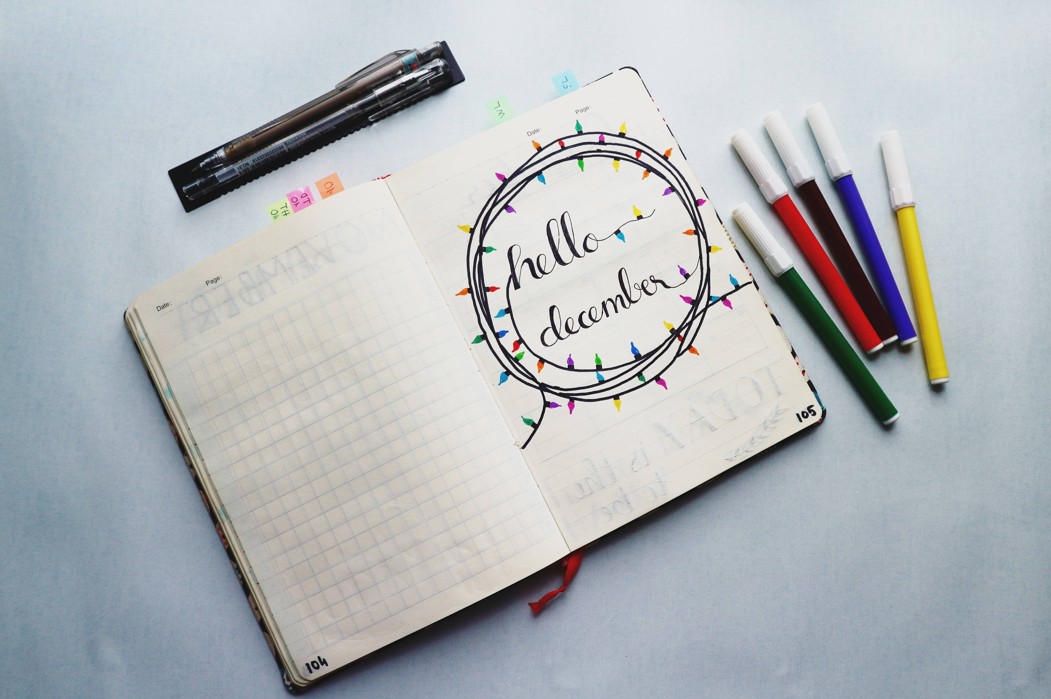 十二月 doodle 日記 壁紙 — 一本打開的筆記本，上面用草書寫著「hello december」，周圍環繞著彩色燈串，旁邊放著一組馬克筆和一支筆，在淺灰色表面上。