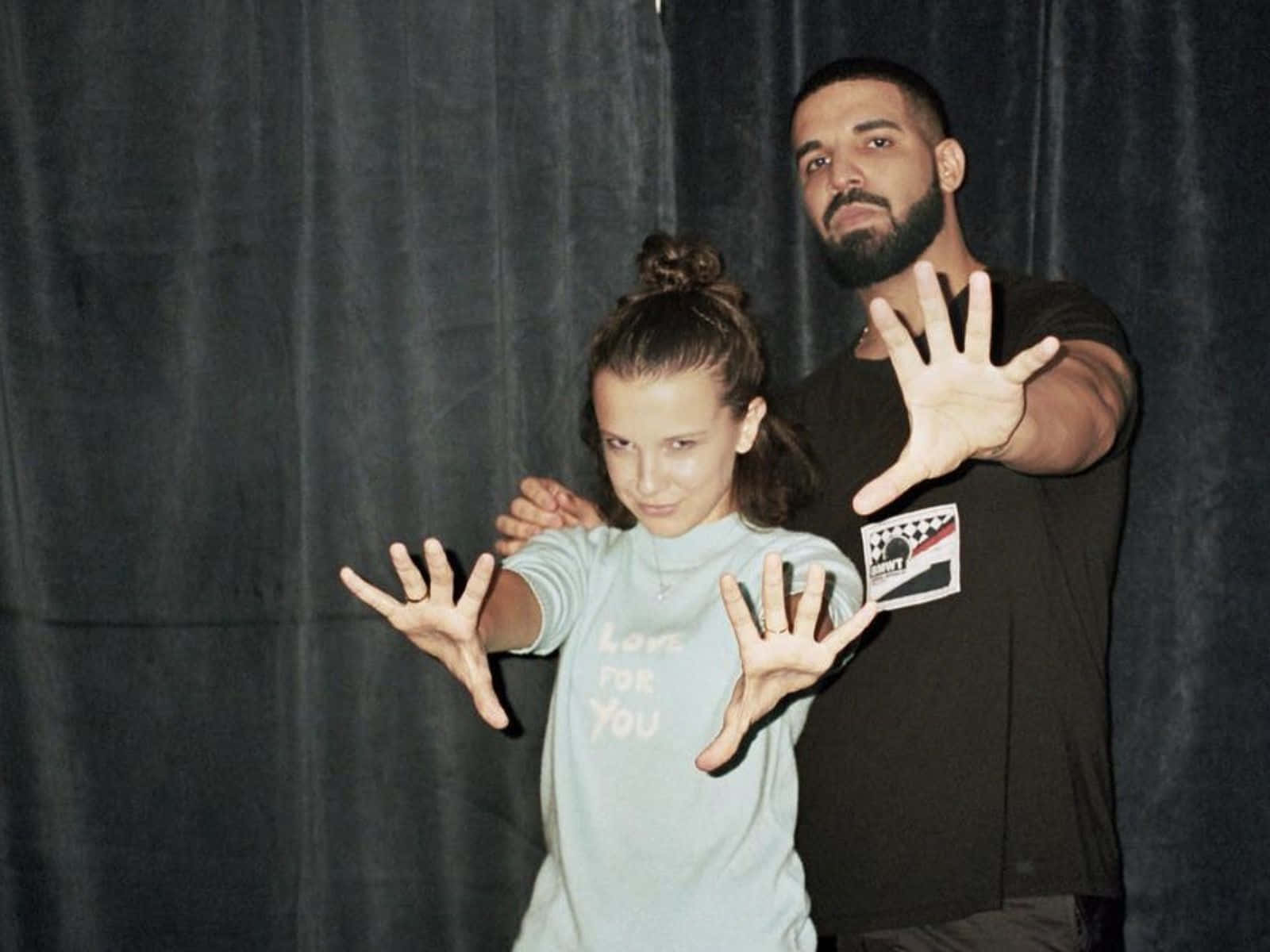 Drake 桌布 壁紙 — Drake 和 Millie Bobby Brown 在深色窗簾背景下伸出手擺姿勢。