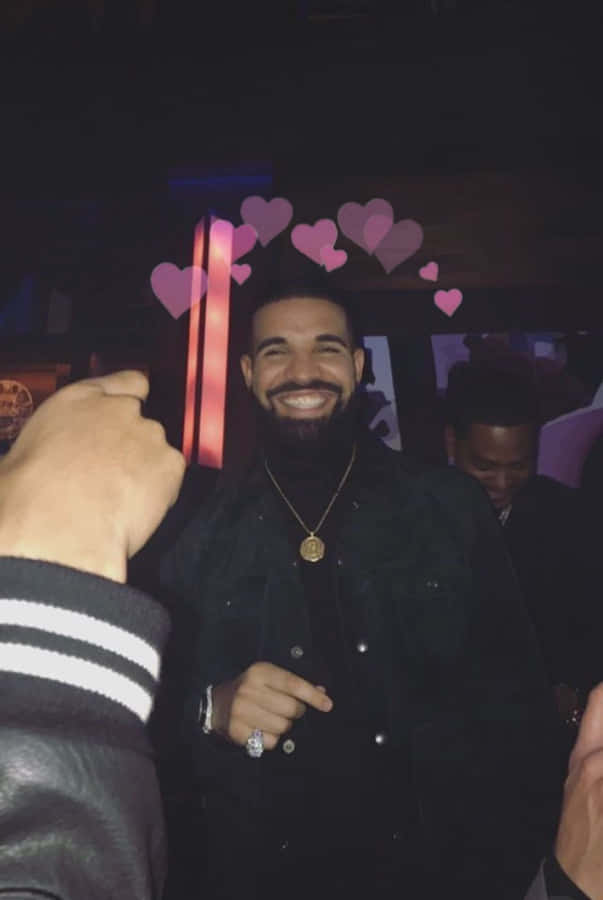「#MoveWithIt」 壁紙 — Drake 在一個燈光昏暗的房間裏的派對上燦爛地微笑，粉紅色的心形圖案漂浮在他的頭頂上方。