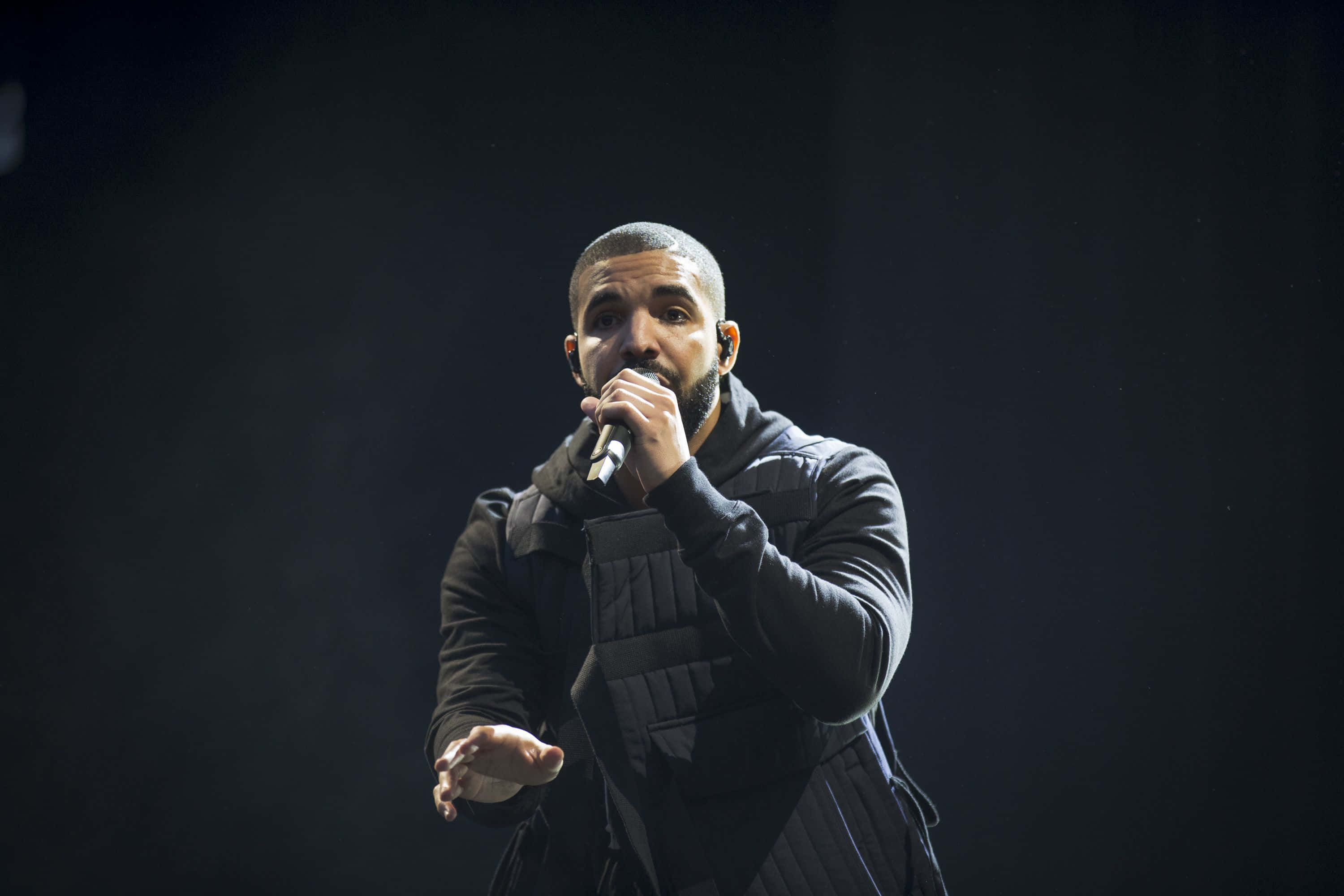 Drake 桌布 壁紙 — Drake 在戲劇性的聚光燈下於舞台上表演，身穿深色加墊外套，表情專注而強烈。