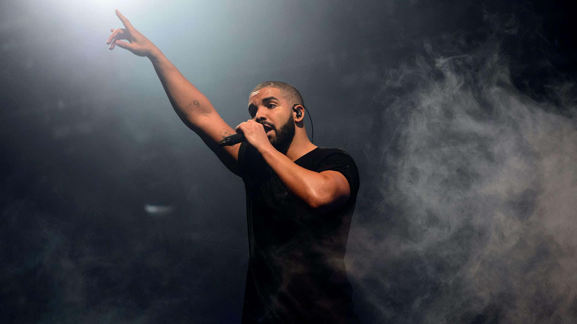 Drake 桌布 壁紙 — Drake 在明亮的聚光燈下，於黑暗煙霧中在舞台上表演，手持麥克風並向上指。