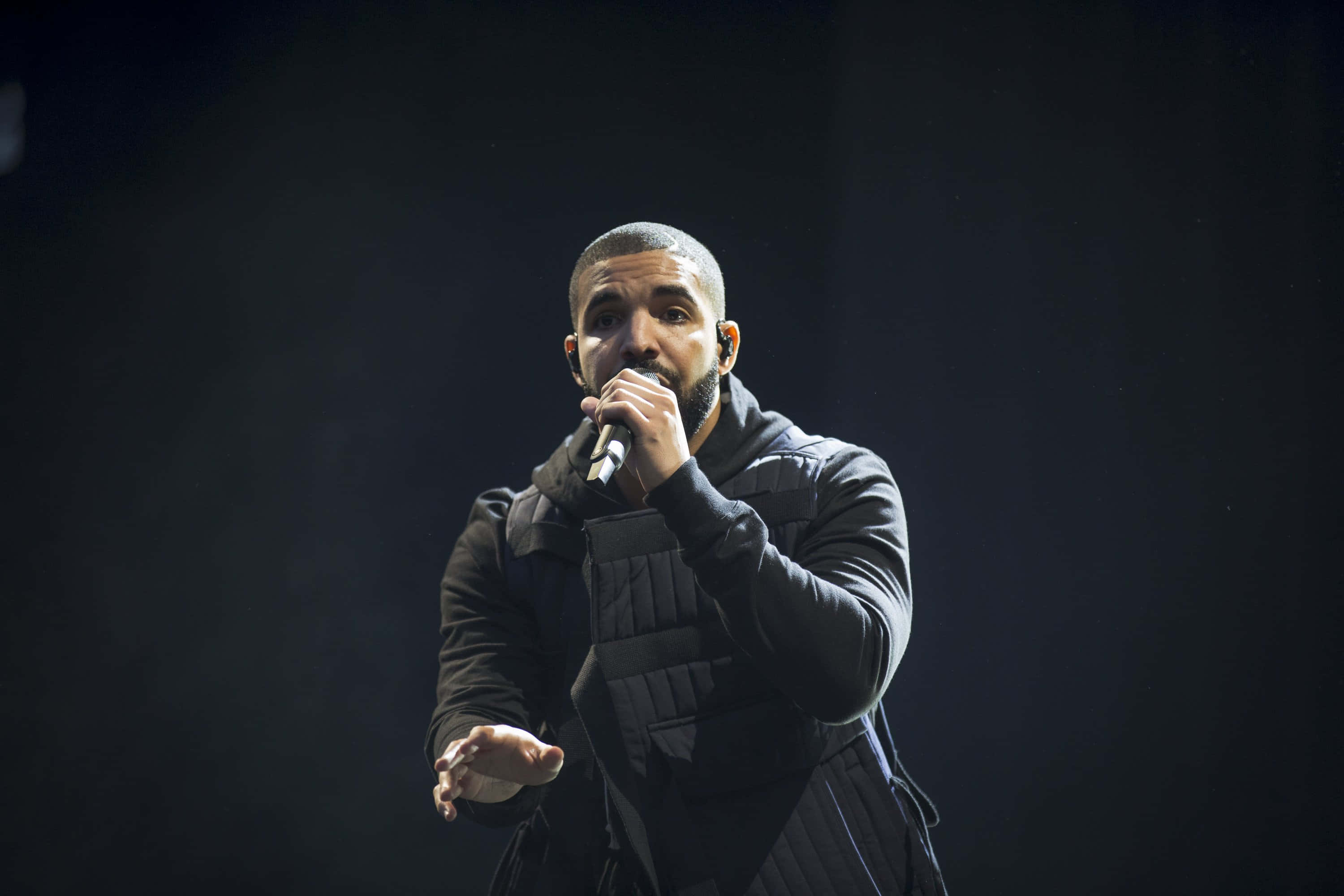 Drake 桌布 壁紙 — Drake 在舞台上穿著深色絎縫外套，在戲劇性的聚光燈下握著麥克風表演。