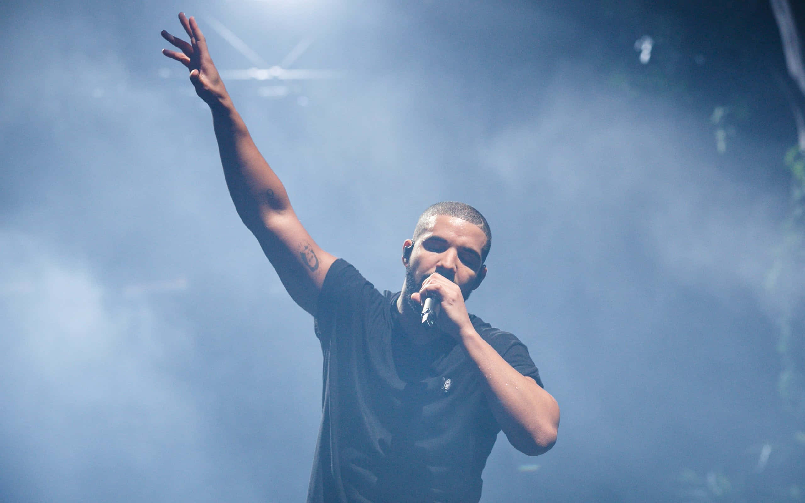 Drake 桌布 壁紙 — Drake 在舞台上表演，在明亮的舞台燈光下，穿過朦朧的藍色煙霧舉起一隻手臂。