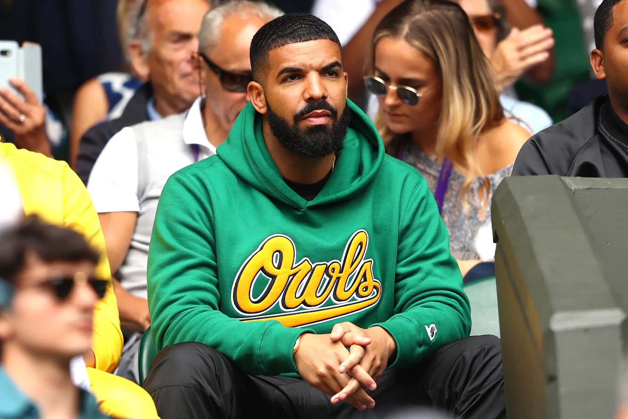 Drake Ovo 桌布 壁紙 — Drake 坐在體育館人群中，穿著綠色 'Owls' 連帽衫，在日間活動中神情專注。