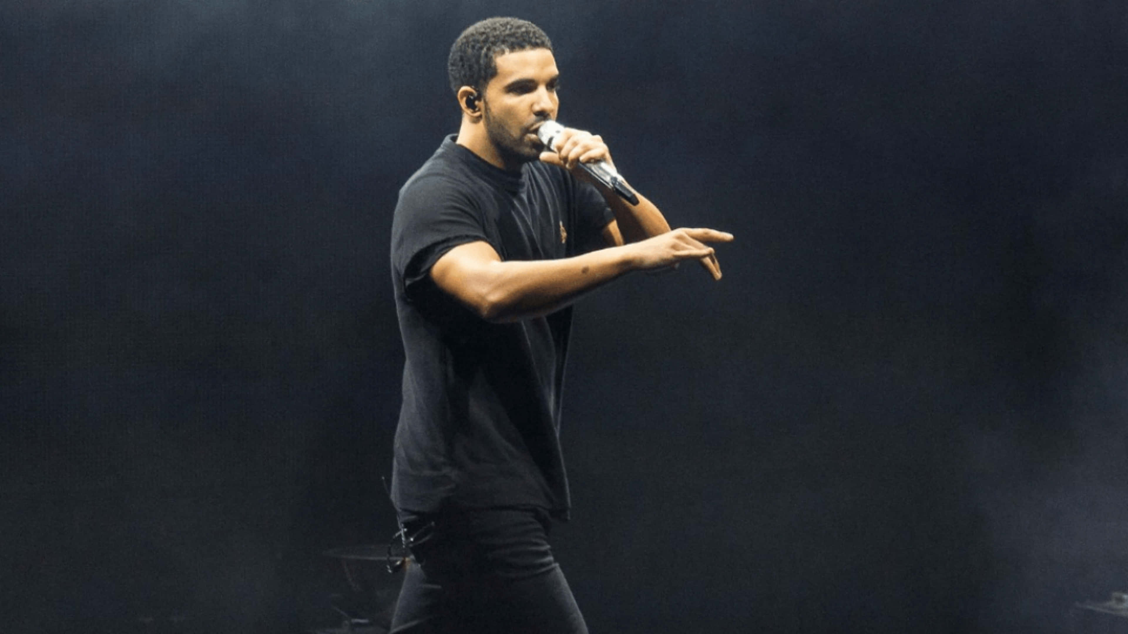 Drake 桌布 壁紙 — Drake 身穿黑色服裝在舞台上表演，在戲劇性的舞台燈光下手持麥克風。