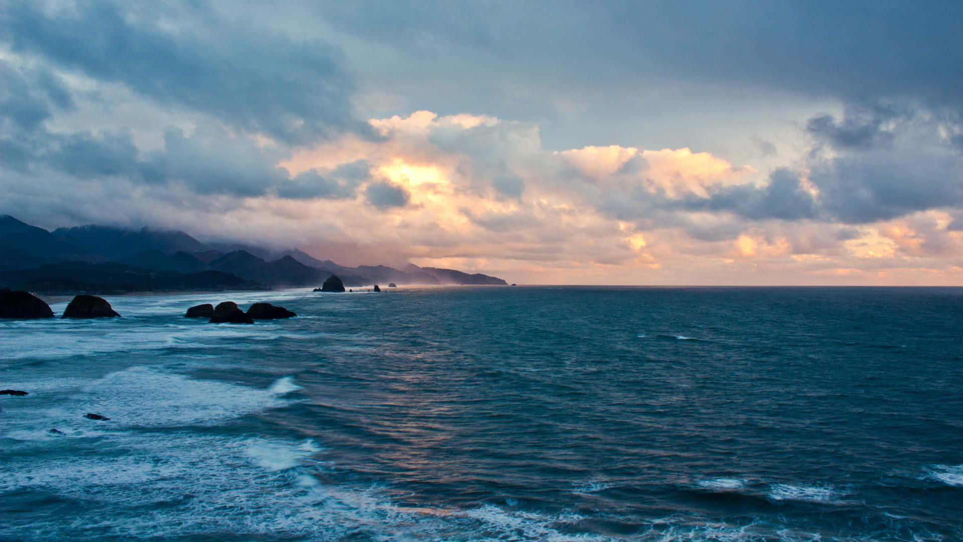 Coastal Sunset 桌布 壁紙 — 戲劇性的海浪在陰沉的黃昏天空下拍打著黑暗的岩石，天空中有發光的雲彩。