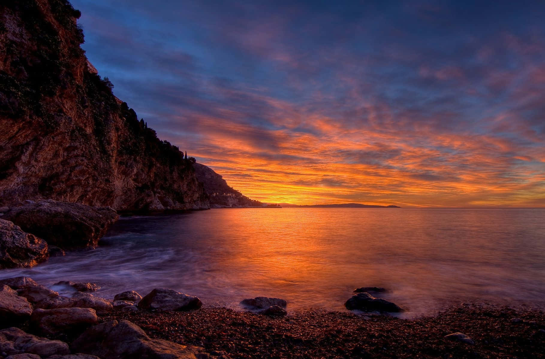 Coastal Sunset 桌布 壁紙 — 戲劇性的日落將天空染成火熱的橙色和深藍色，映照在崎嶇懸崖旁的平靜海面上。