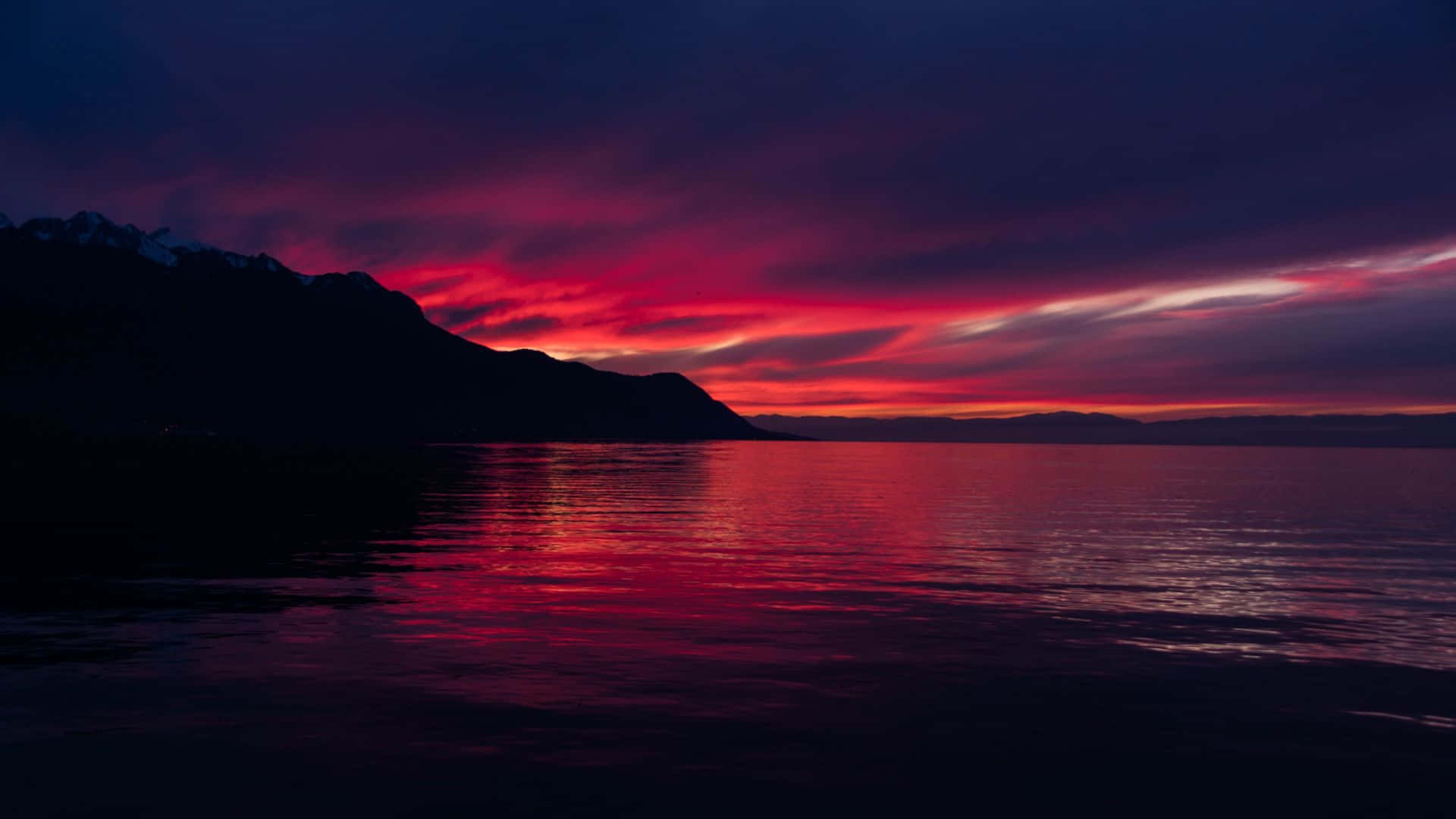 Coastal Sunset 桌布 壁紙 — 戲劇性的黃昏天空，以火紅和深紫色繪成，在平靜的水面上與黑暗的山脈輪廓形成鮮明倒影。