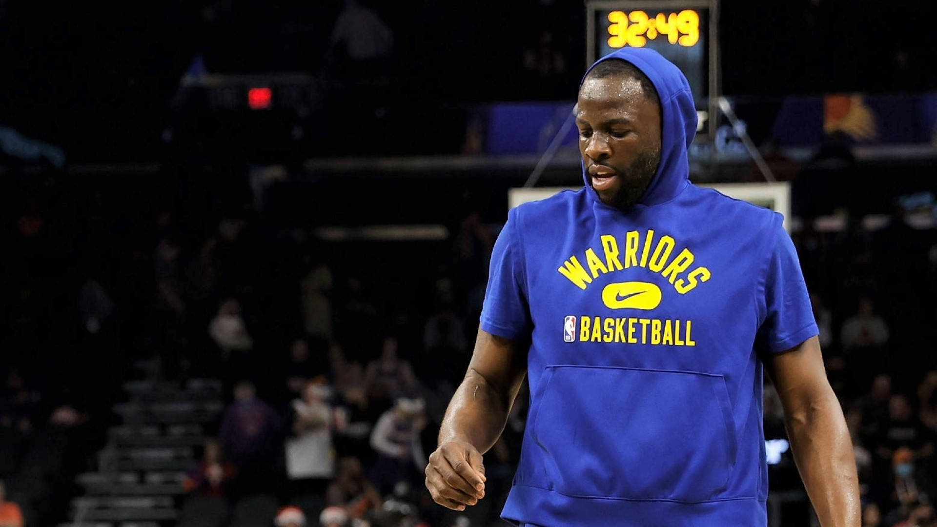 Draymond Green 藍色連帽衫賽前暖身照 壁紙 — 一名身穿藍色勇士隊連帽衫的籃球運動員在球場上行走，背景有顯示32:49的記分牌。