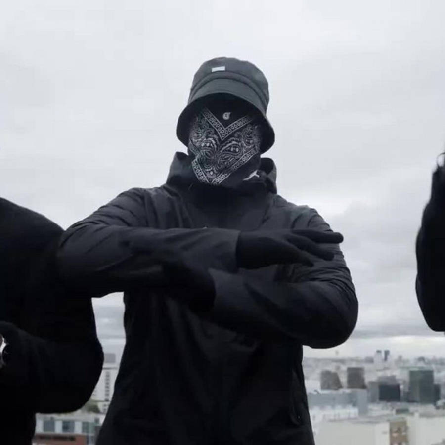 英國Drill Ziak 壁紙 — 一個全身黑色、戴著圖案頭巾和漁夫帽的人，站在一個可以俯瞰城市的天台上，天空多雲。