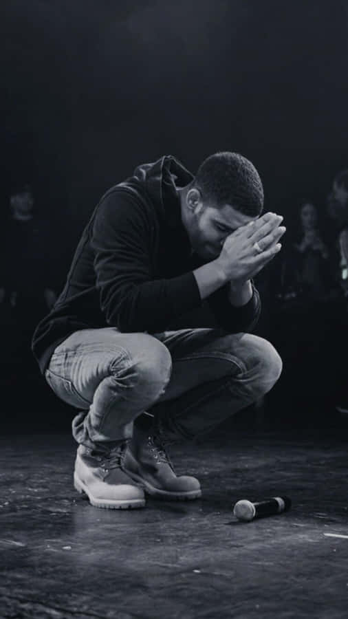 「另一種Drizzy」 壁紙 — 一個男人跪在黑暗的舞台上，雙手掩面，一支麥克風被遺棄在他身旁。