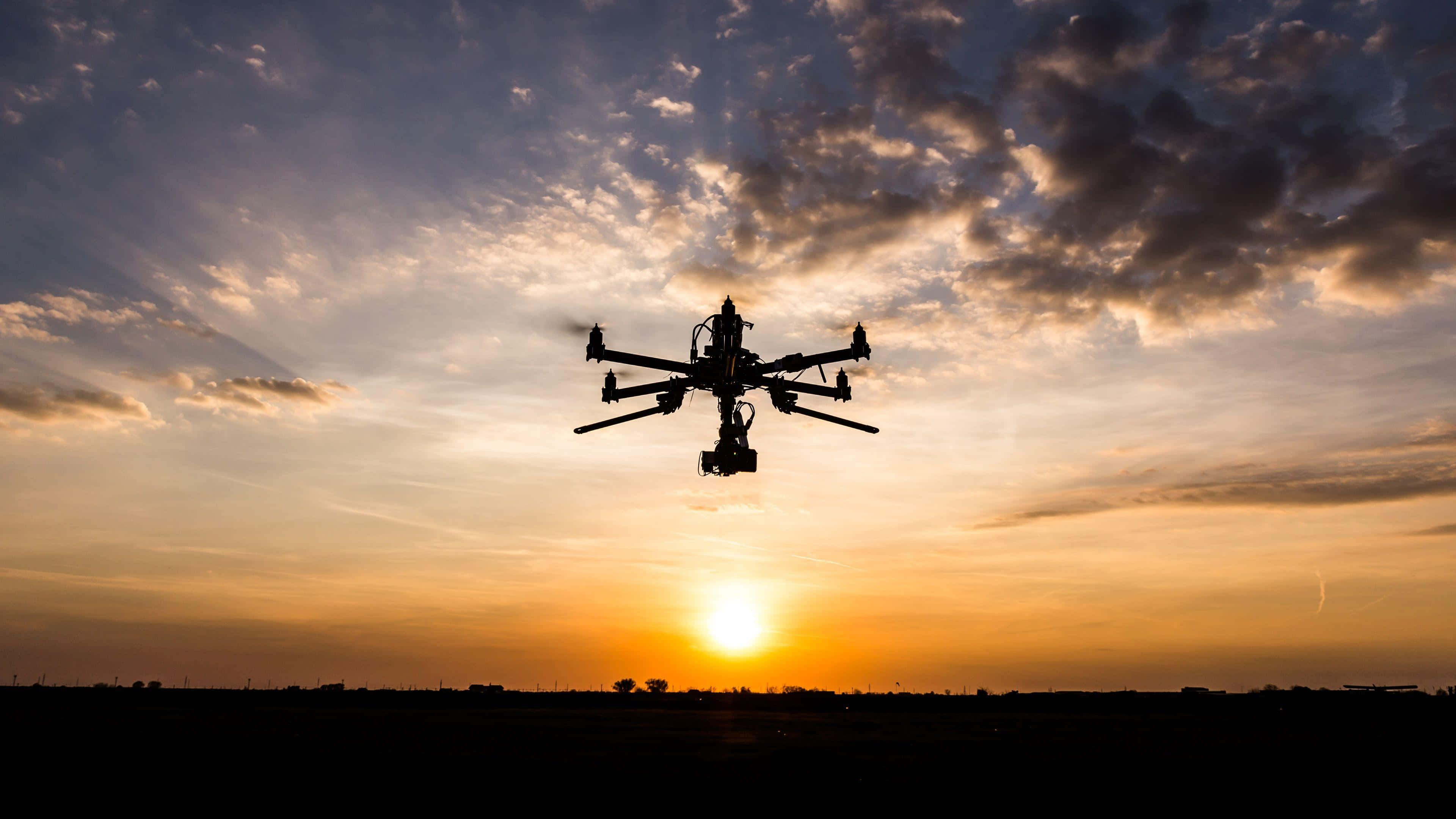 Drone Camera 桌布 壁紙 — 無人機在充滿活力的日落天空背景下呈現剪影，天空中有金色的雲朵和地平線上發光的太陽。