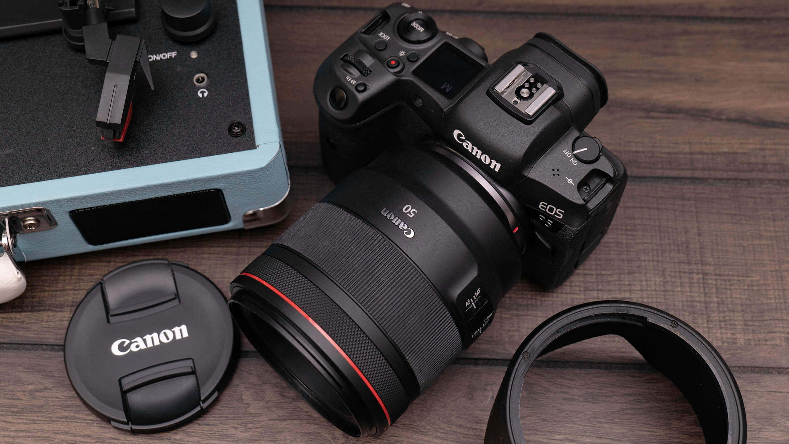 木桌上放置的佳能DSLR相機 壁紙 — 一部黑色Canon EOS R5相機配50mm鏡頭放在木質表面上，旁邊有鏡頭蓋和淺藍色相機袋。