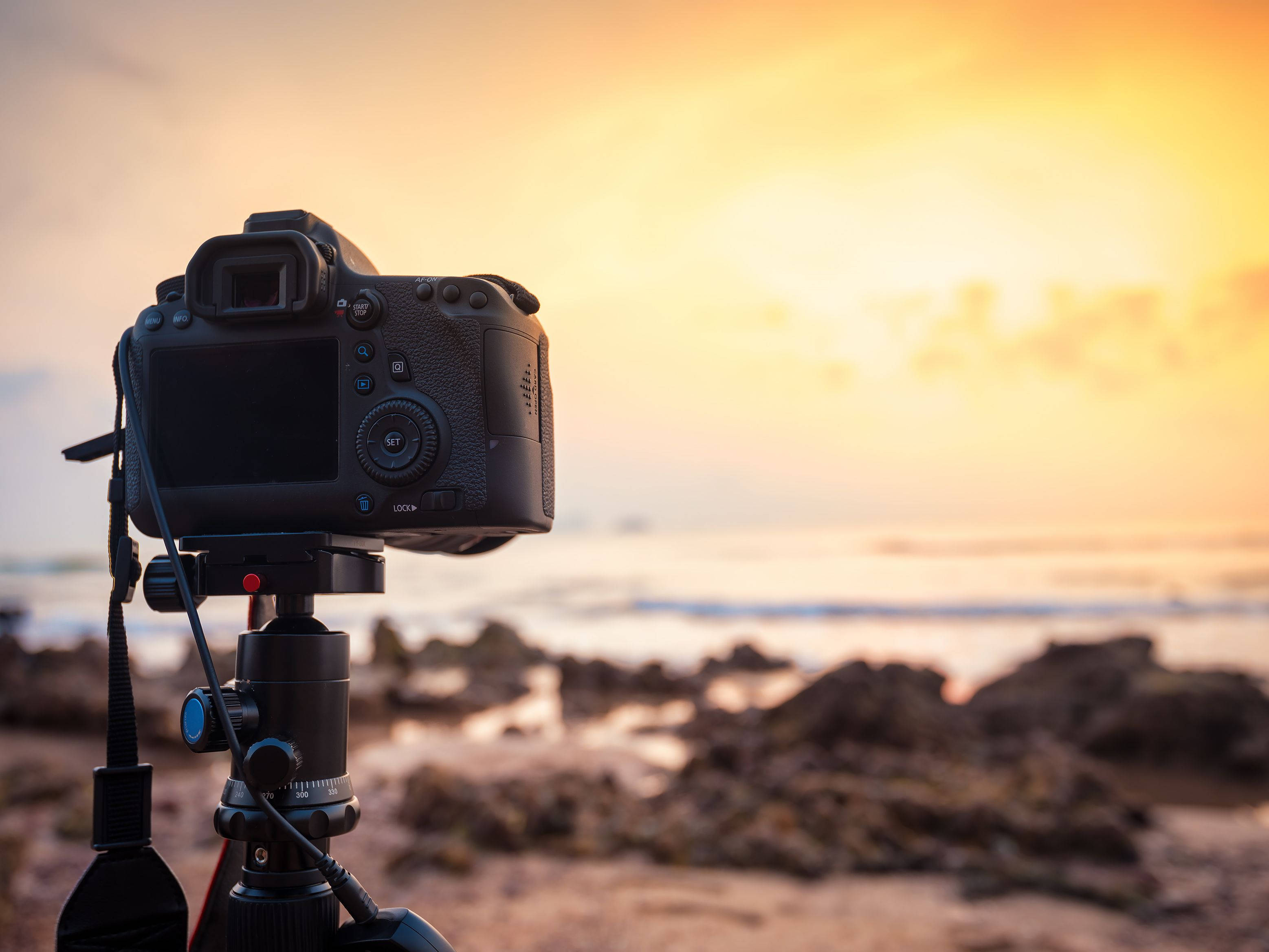 DSLR 相機海景 壁紙 — 安裝在三腳架上的DSLR相機捕捉到岩石海岸線上寧靜的日落，伴隨著溫暖的金色光芒。
