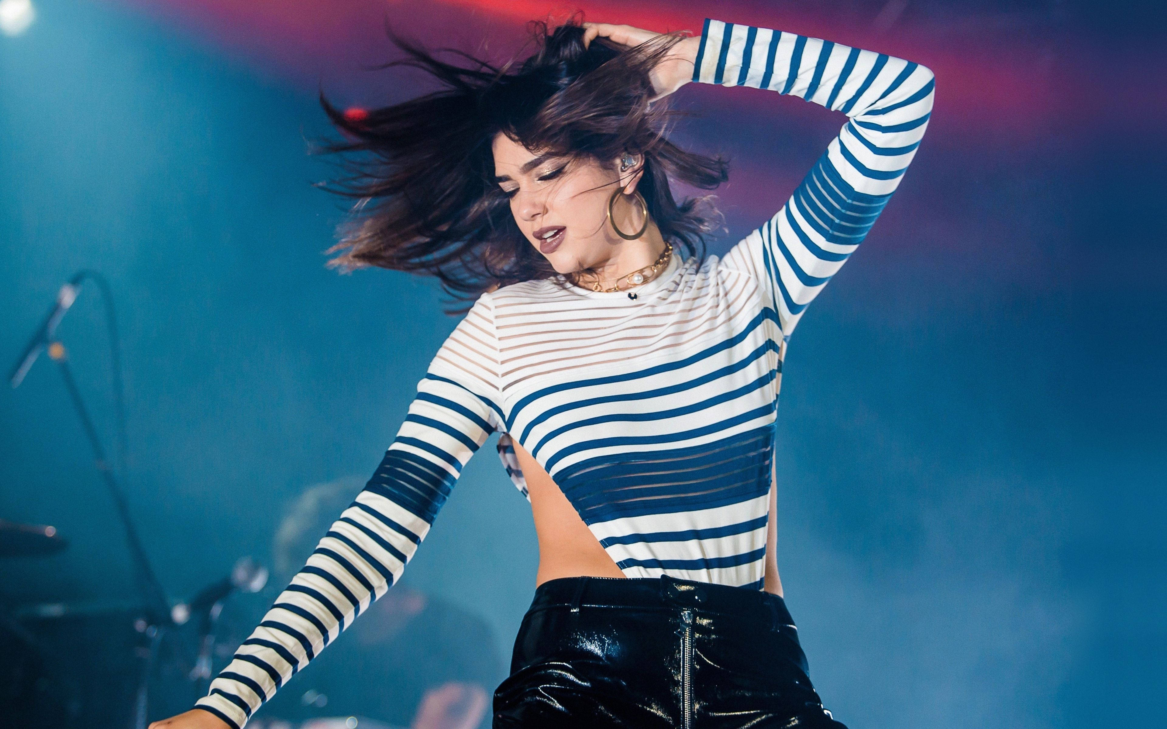 Dua Lipa 4K 巴西演出 壁紙 — 一位充滿活力的女性表演者，頭髮飛揚，身穿條紋上衣，在鮮豔的藍色和紅色舞台燈光下跳舞。