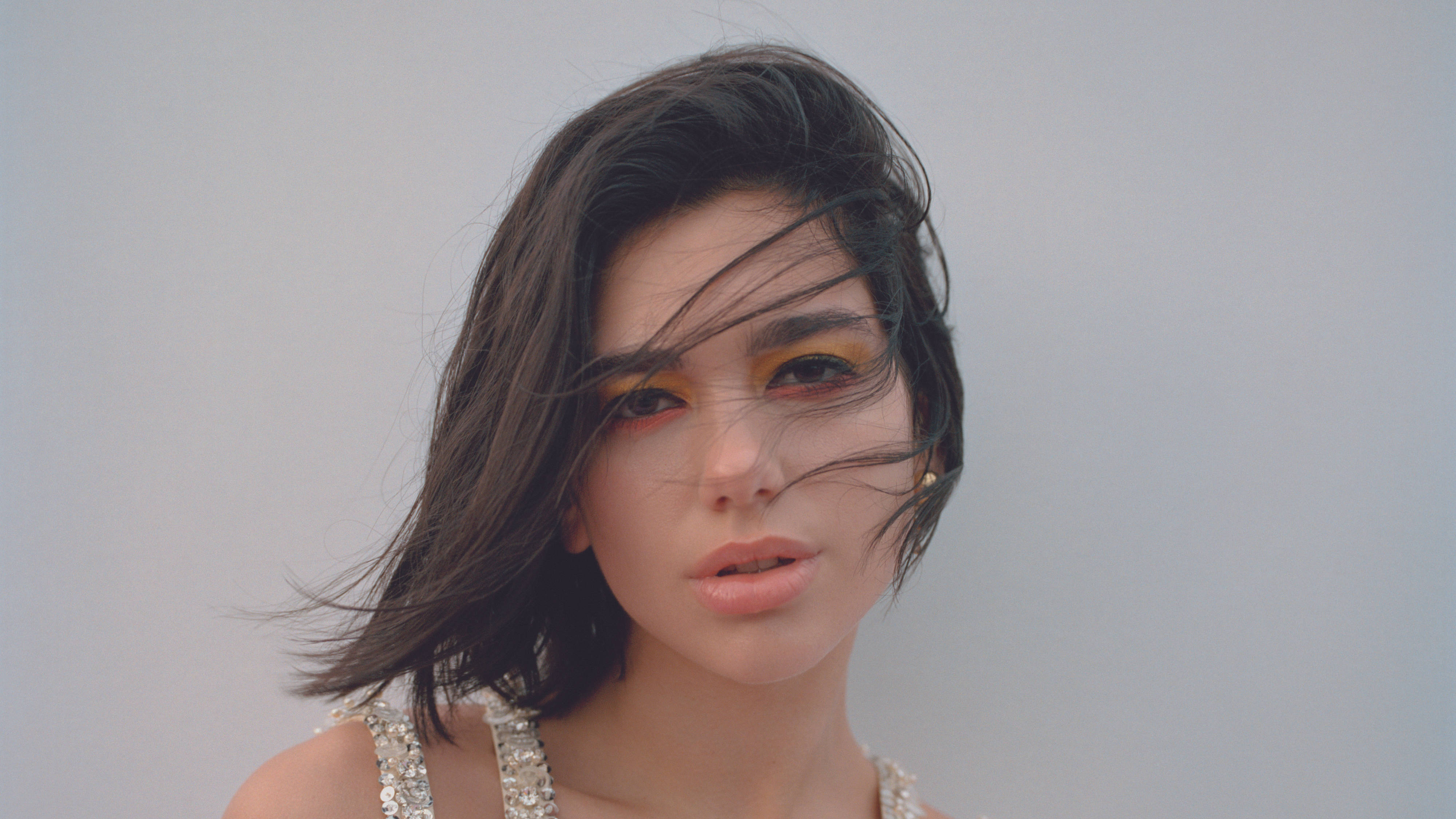 Dua Lipa 精緻妝容 壁紙 — 一位擁有被風吹拂的深色頭髮和大膽黃色眼妝的女性，在柔和的灰色背景下直視鏡頭。