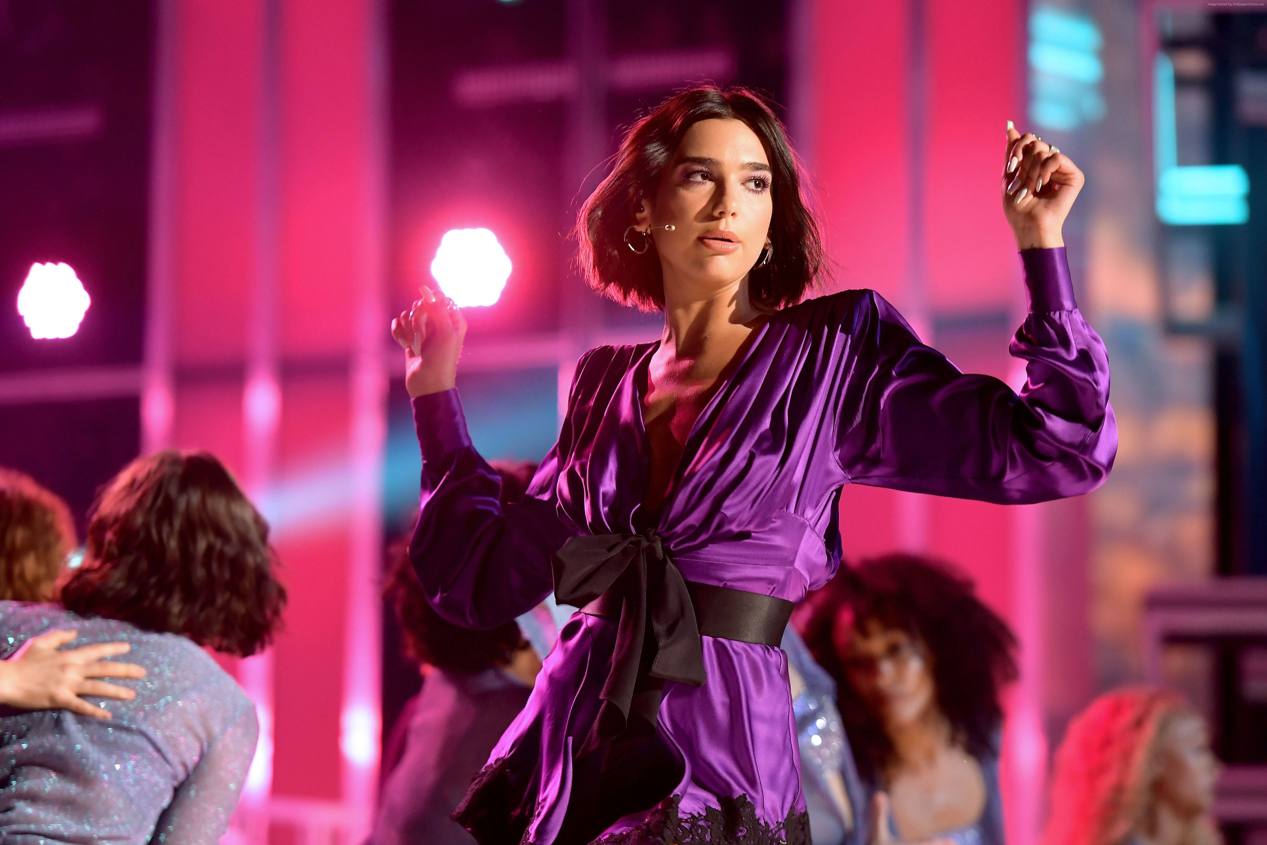 Dua Lipa 紫色禮服 壁紙 — 一位身穿閃亮紫色緞面連衣裙的女性在充滿活力的粉紅色舞台燈光下擺出自信的姿勢。
