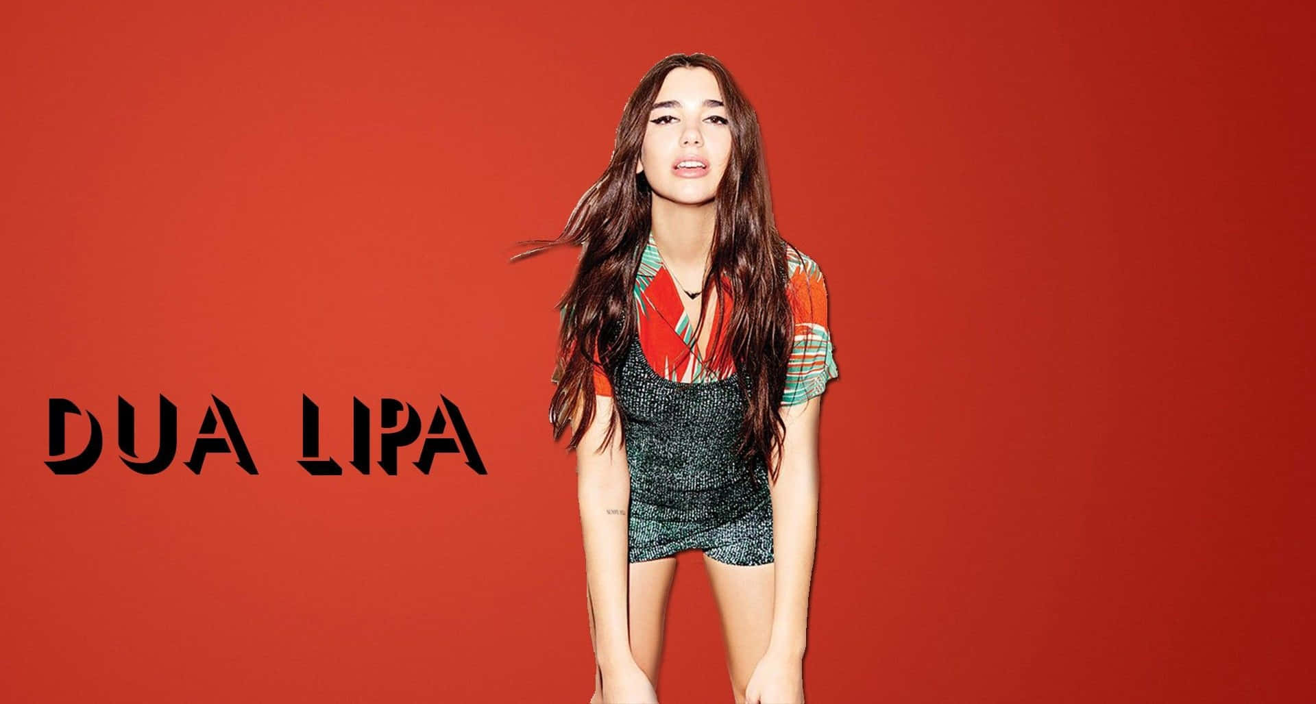 杜娃·利帕 桌布 壁紙 — Dua Lipa 自信地站在大膽的紅色背景前，身穿彩色圖案上衣和閃亮短褲。