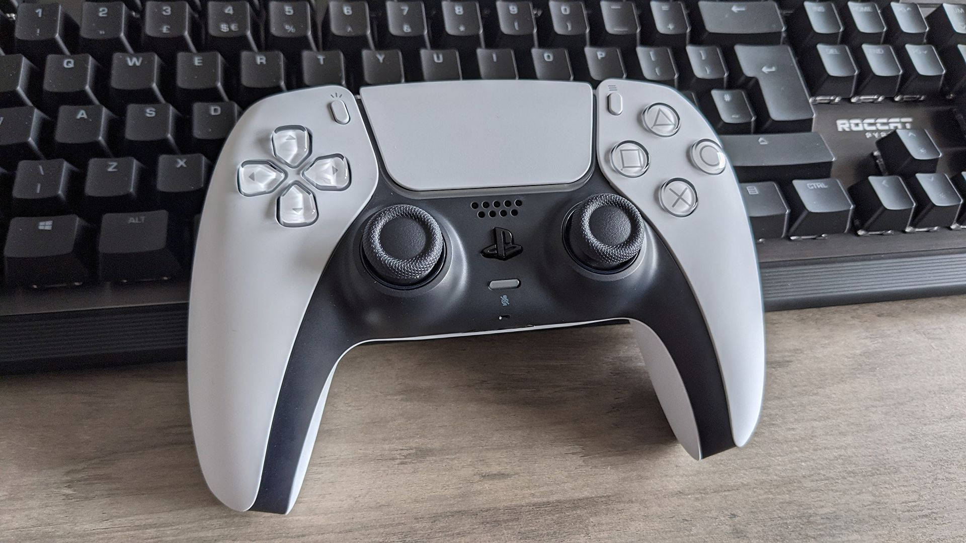 DualSense PS5控制器 壁紙 — 一個時尚的銀黑色PlayStation 5 DualSense控制器放在木桌上，旁邊有一個黑色鍵盤。