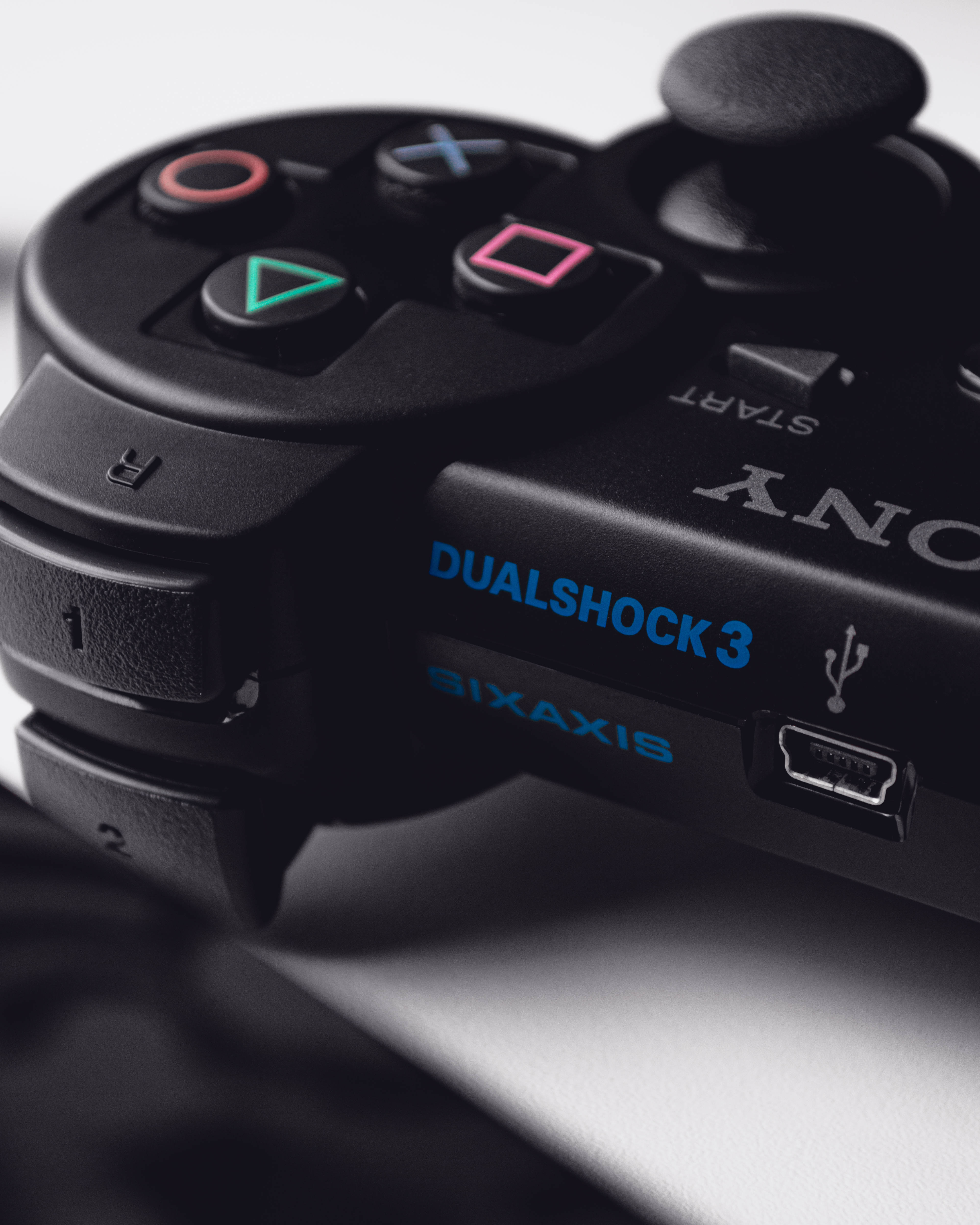 索尼 DualShock PS3 控制器 壁紙 — 黑色PlayStation DualShock 3手掣的特寫，帶有發光的彩色按鈕和藍色文字，置於柔軟的白色表面上。