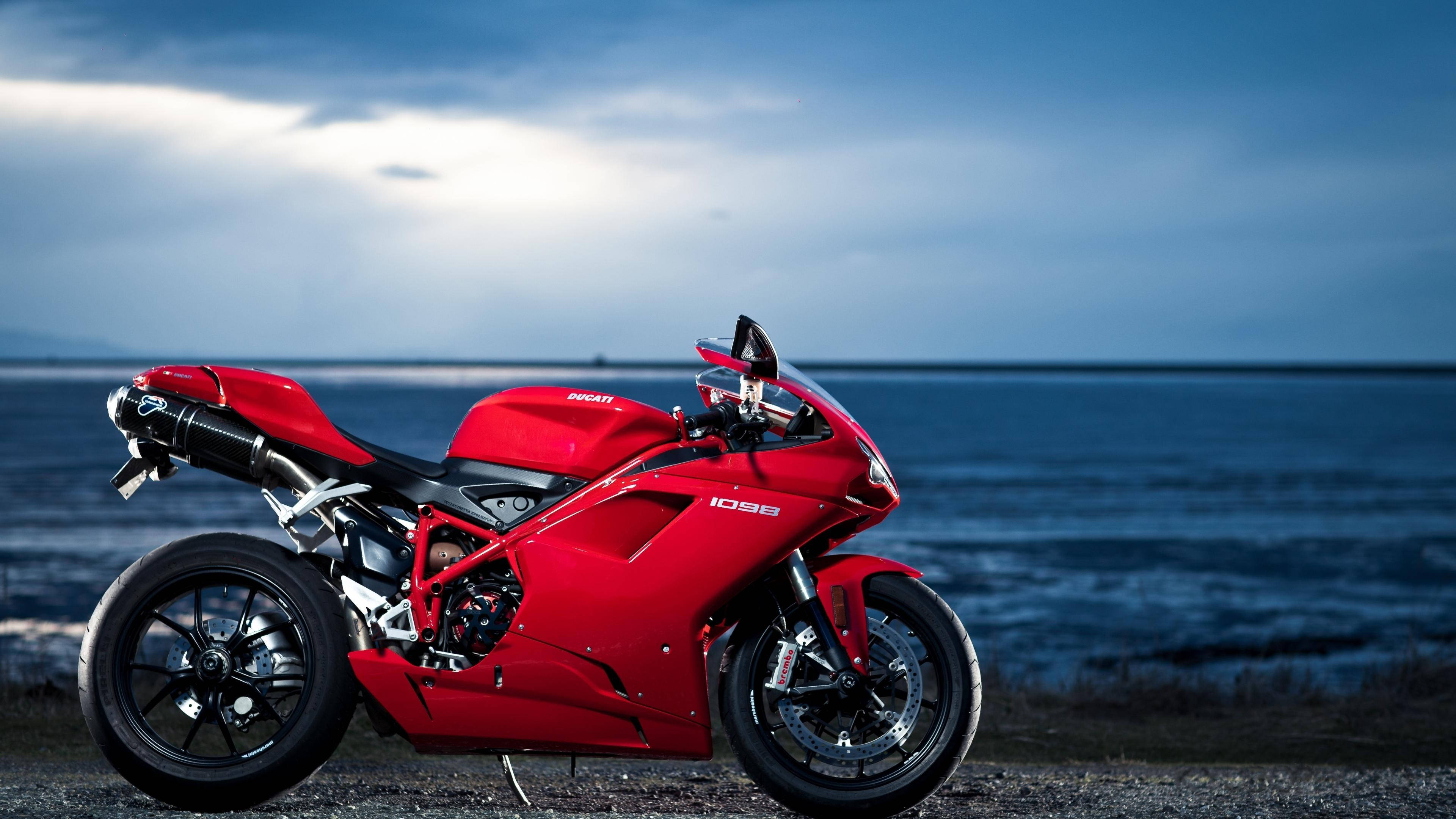 Ducati 4K摩托車 壁紙 — 一架搶眼的紅色Ducati 1098電單車，在暮色海洋和密雲天空的背景下，佇立在碎石岸邊。