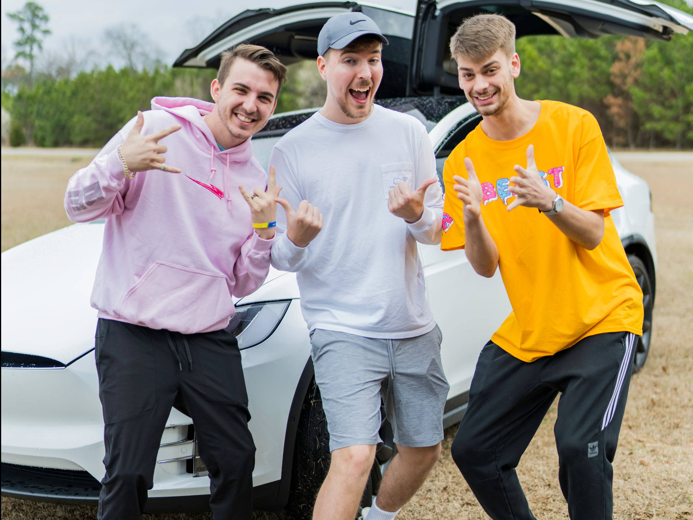 Dude Perfect 三人與特斯拉Model X 壁紙 — 三位身穿色彩繽紛街頭服飾的年輕男子，在草地上的一輛白色Tesla SUV前充滿活力地擺姿勢。